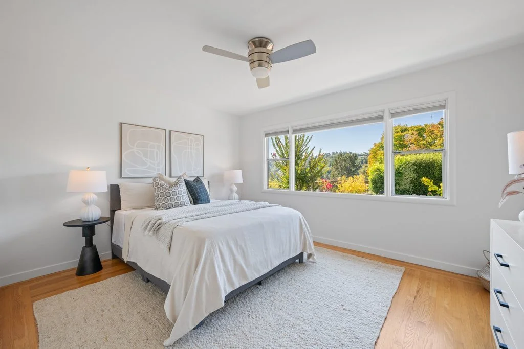 Bright bedroom with white walls, a queen-sized bed with white bedding and decorative pillows, two white lamps on black nightstands, a ceiling fan, and large windows showing an outdoor view of trees and blue sky.