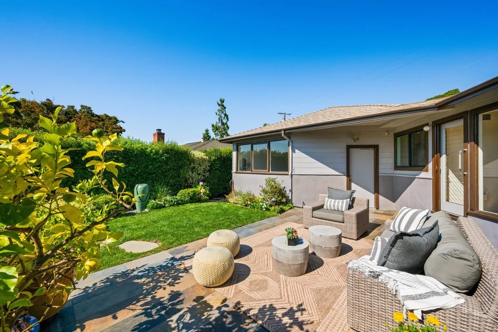 A bright backyard patio with outdoor furniture, including a wicker sofa and chairs with cushions, circular side tables, and a beige outdoor rug. The patio is adjacent to a house with sliding doors, and the yard features a green lawn, shrubs, and a he