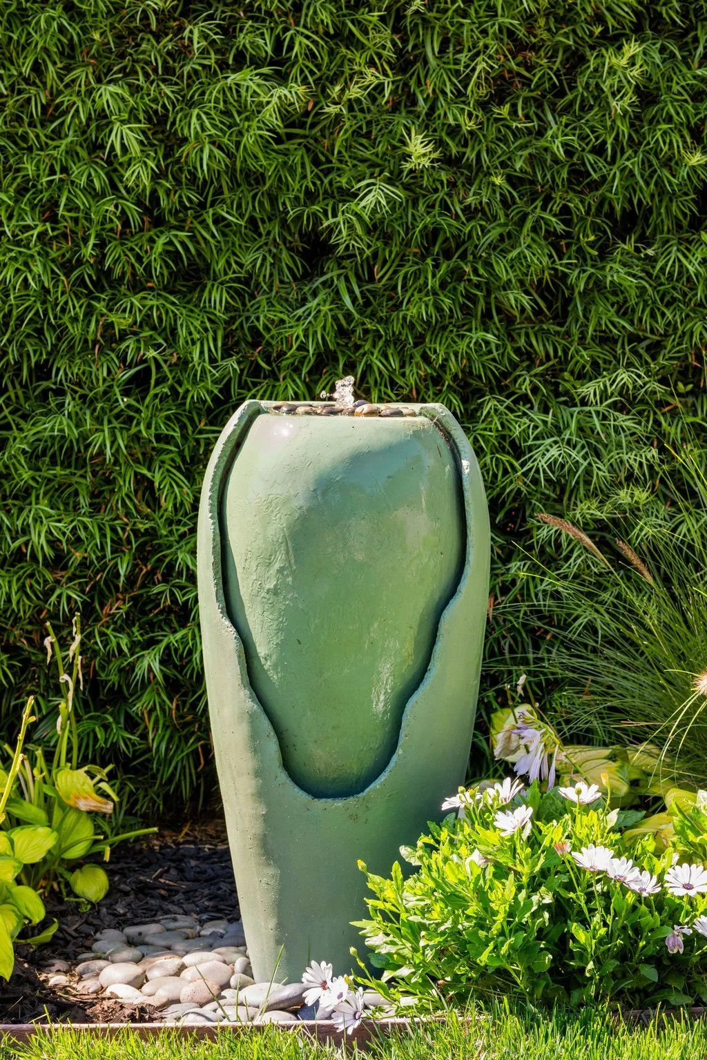 Outdoor garden scene with a modern green water fountain surrounded by flowers and lush green plants.