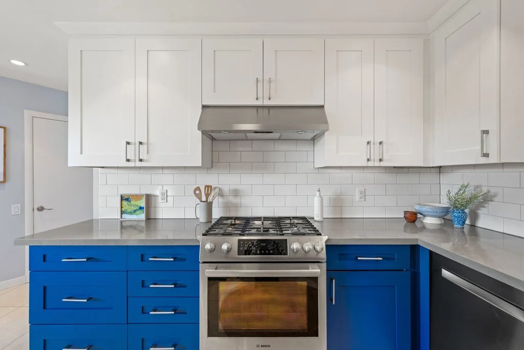 Modern kitchen with white upper cabinets, blue lower cabinets, stainless steel stove with oven, white subway tile backsplash, gray countertop, and various kitchen items including utensils, a soap dispenser, and decorative bowls and plants.