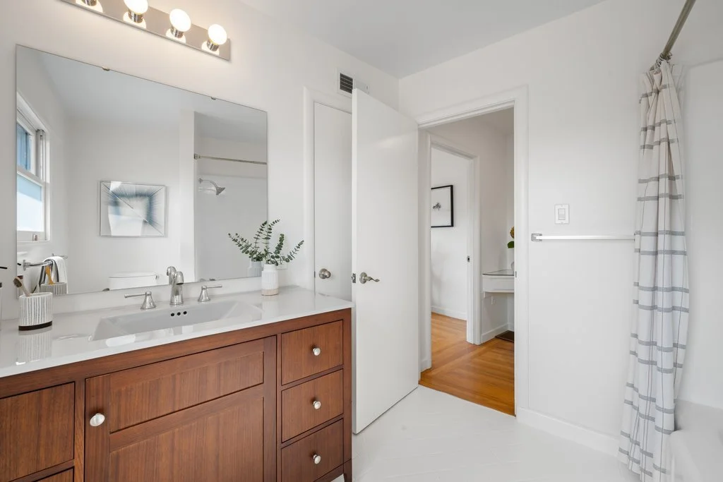 Bathroom vanity with a mirror, a plant, and bathroom accessories, with a partially open door leading to a hallway and a view of a shower area and another room.