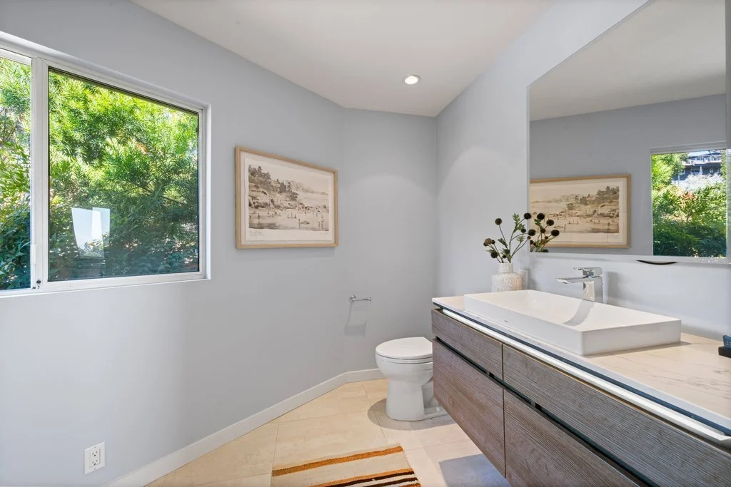Modern bathroom with a white vessel sink, wooden vanity, framed artwork, and a large mirror, with two windows showing greenery outside.