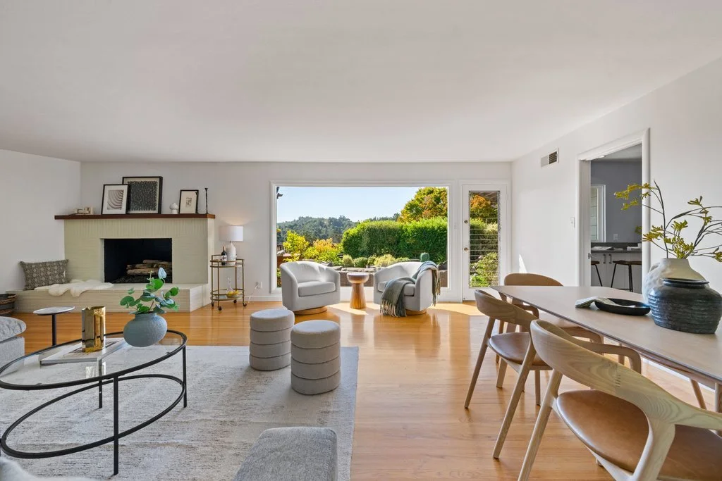 Bright living room with white walls, hardwood floors, and large windows showing outdoor greenery. Decor includes a fireplace, chairs, a coffee table, and a dining table with chairs.