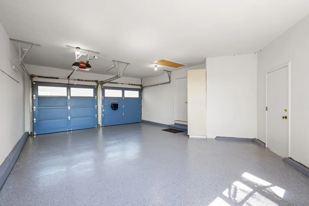 Empty garage with blue sectional doors, gray speckled floor, white walls, and a small staircase at the back.