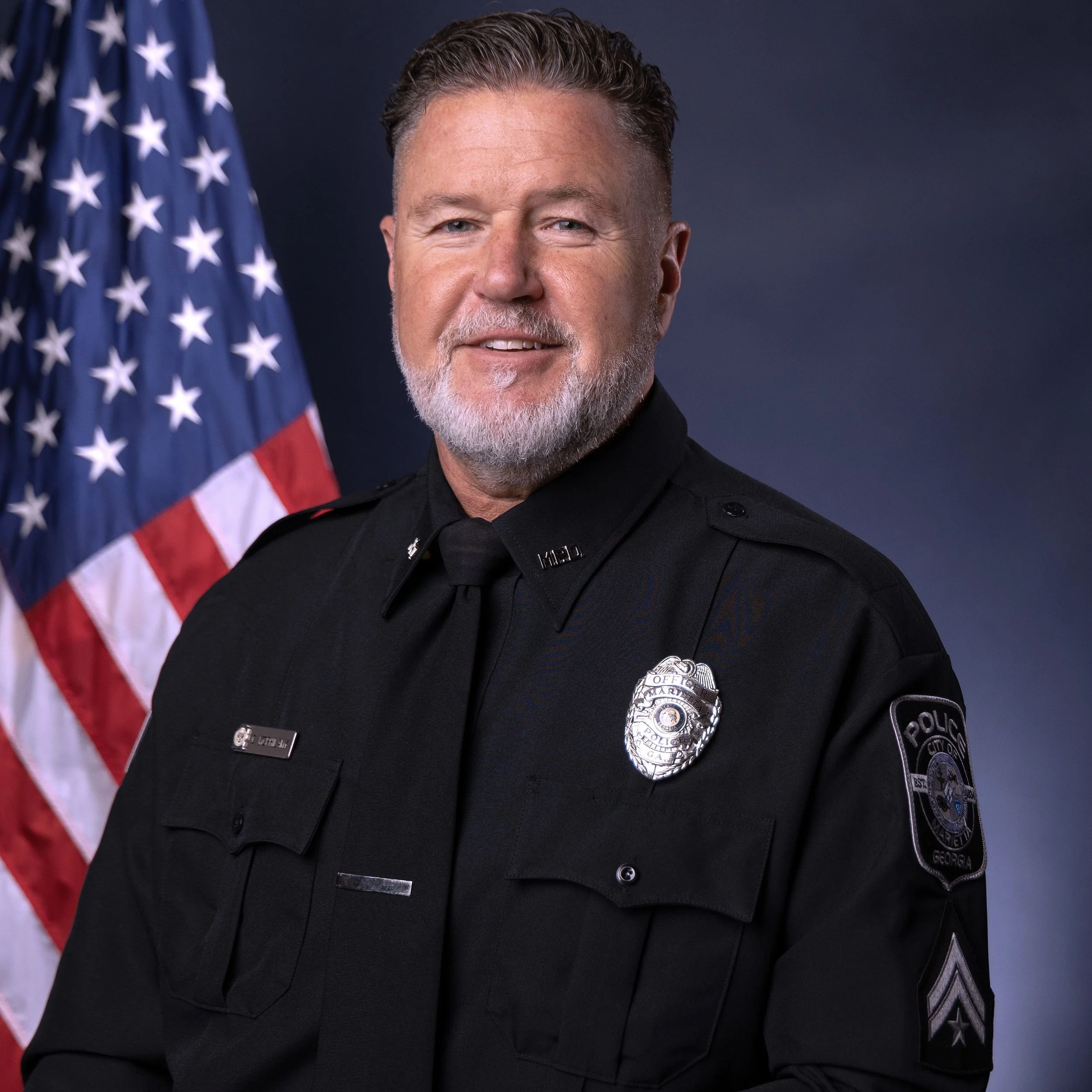 Close-up of a male police officer with gray beard and short hair, wearing a black police uniform with a badge, standing in front of an American flag.