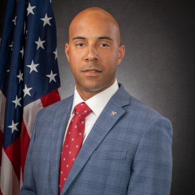 A man in a blue suit with a red tie standing in front of an American flag.