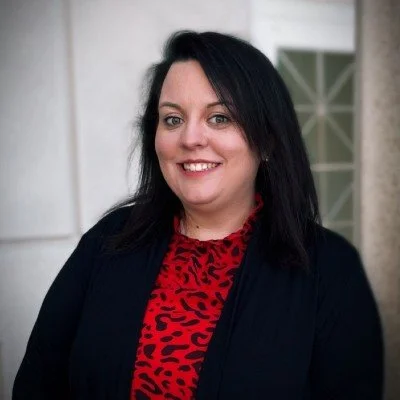A woman with black hair wearing a red patterned blouse and a black blazer, smiling in front of a white building with windows.