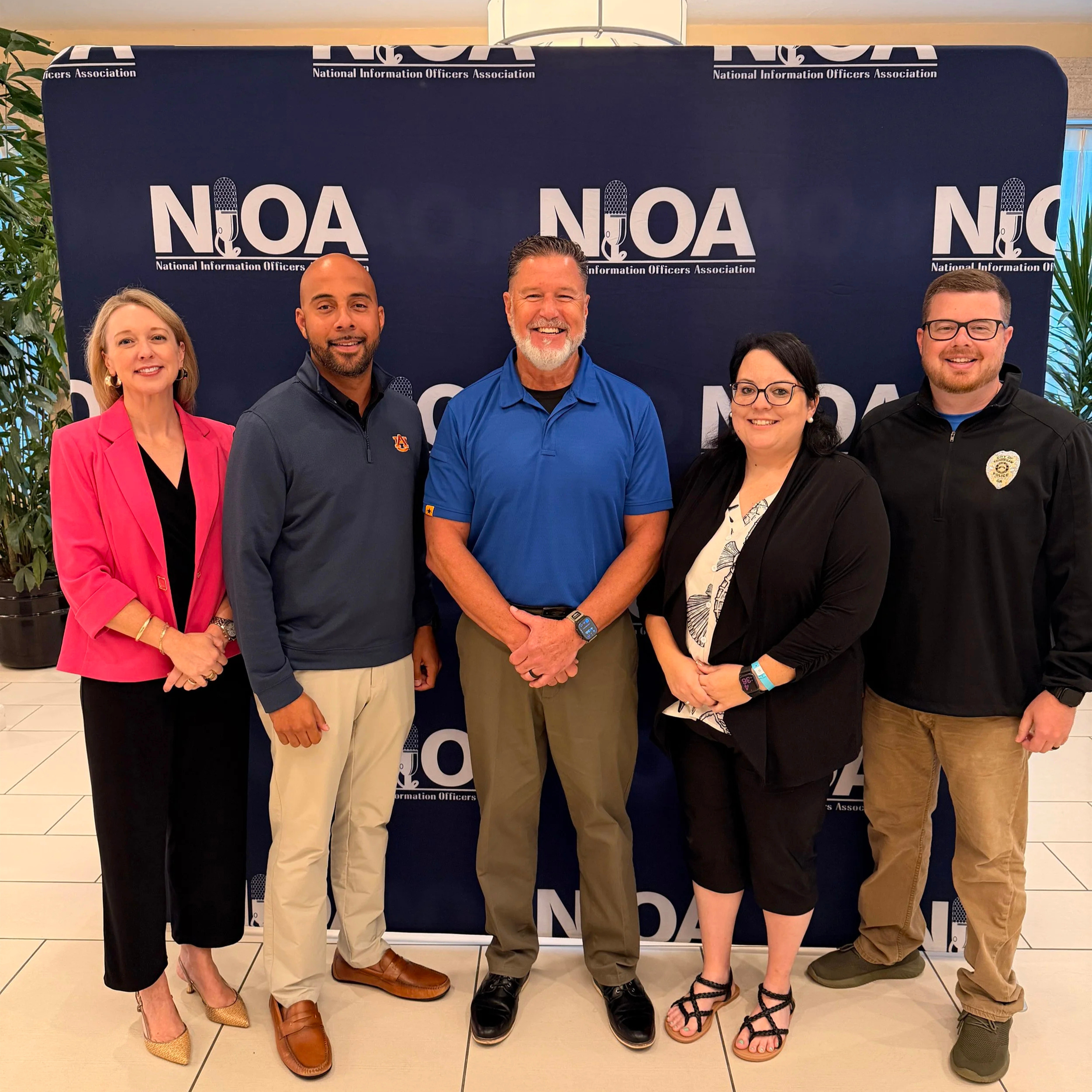Group of five people standing together in front of a blue backdrop with the logo of the National Information Officers Association. They are dressed in business casual clothing and smiling at the camera.
