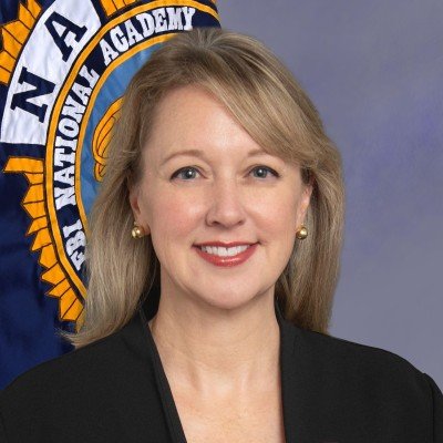 A woman with blonde hair, wearing a black top and pearl earrings, smiling in front of an FAA emblem.