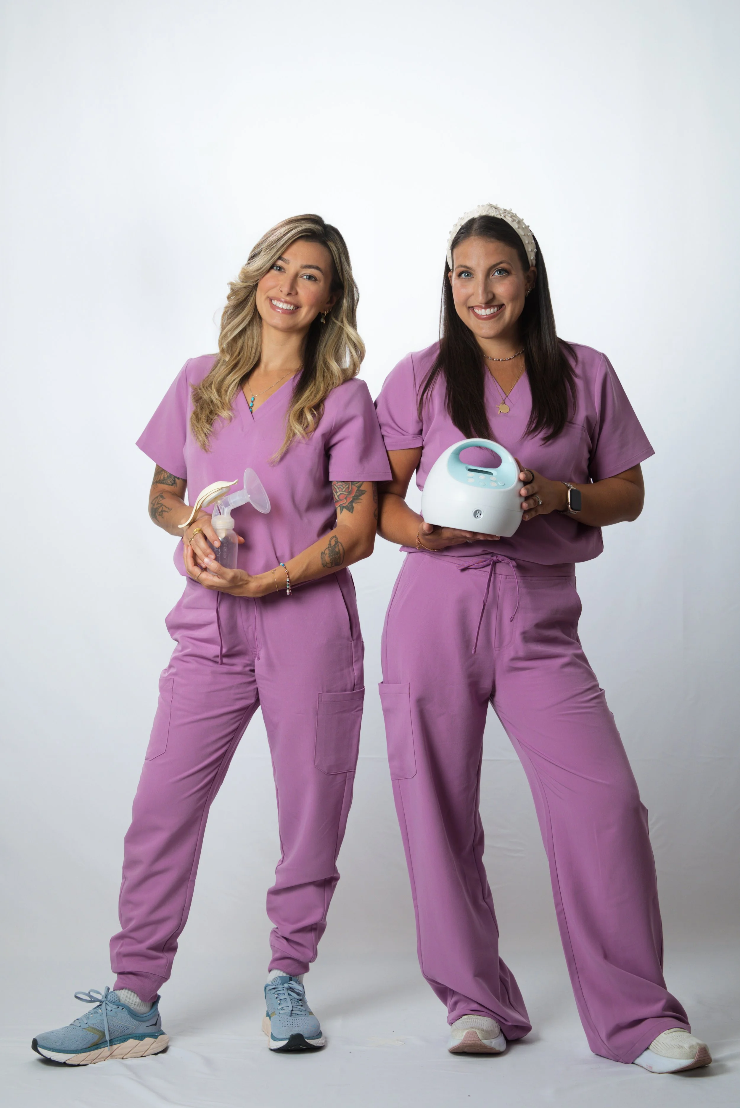 Two women dressed in purple scrubs standing side by side against a plain white background. The woman on the left holds a spray bottle, and the woman on the right holds a small device. Both women are smiling.