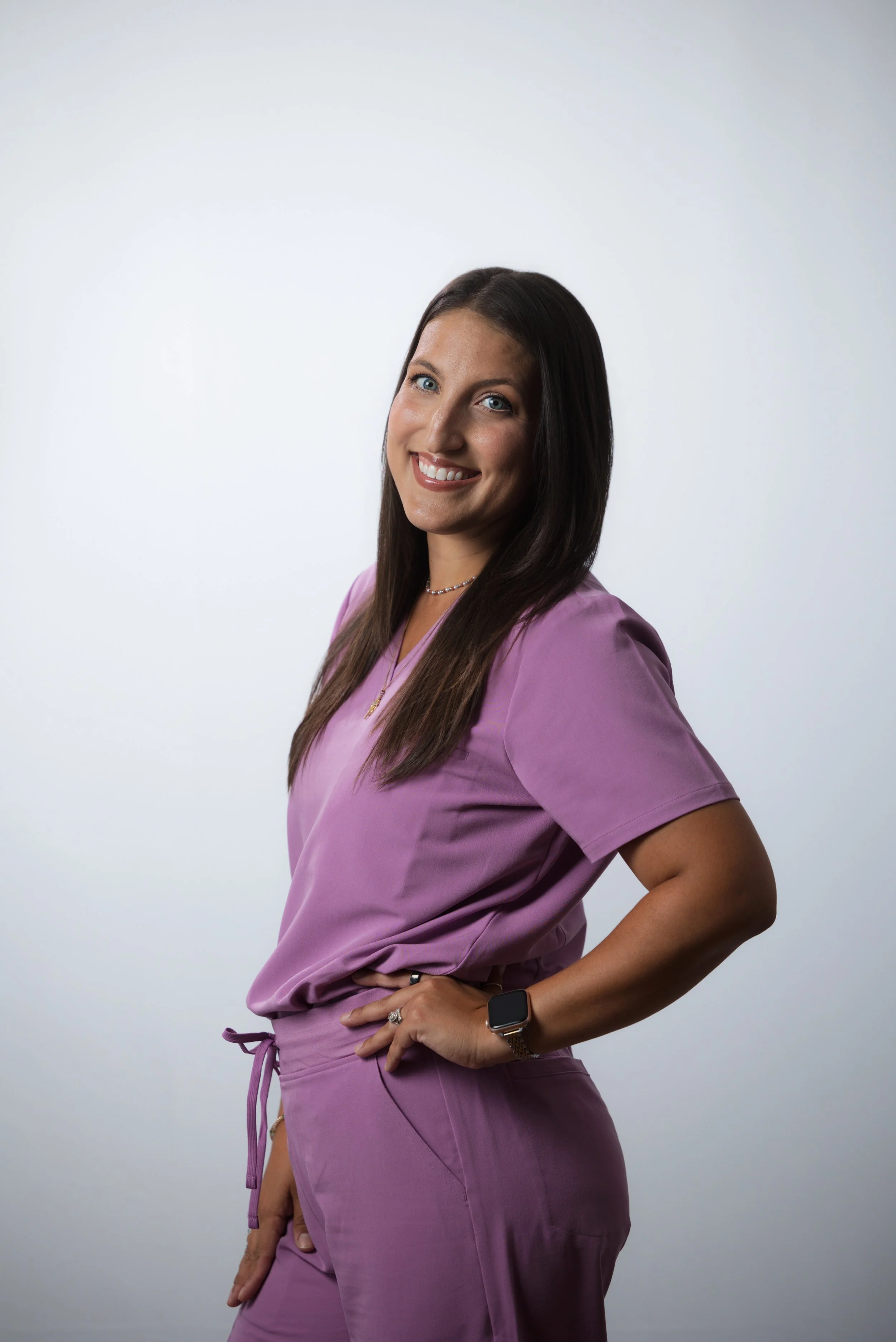 A woman in purple scrubs with long dark hair, smiling and posing with one hand on her hip against a plain light background.