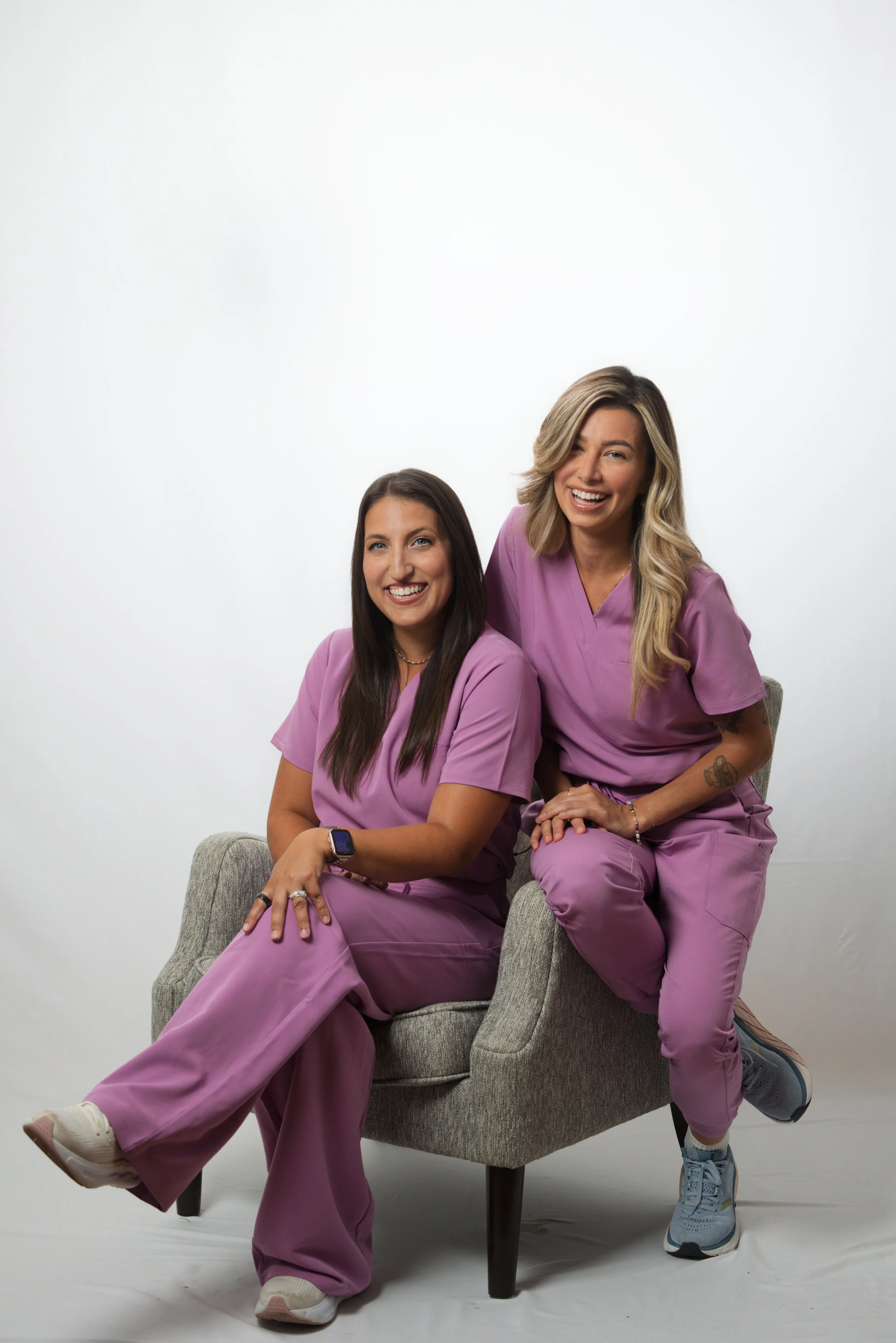 Two women wearing medical scrubs sitting on and leaning against a gray chair in a studio with a plain white background. They are smiling and appear to be happy.
