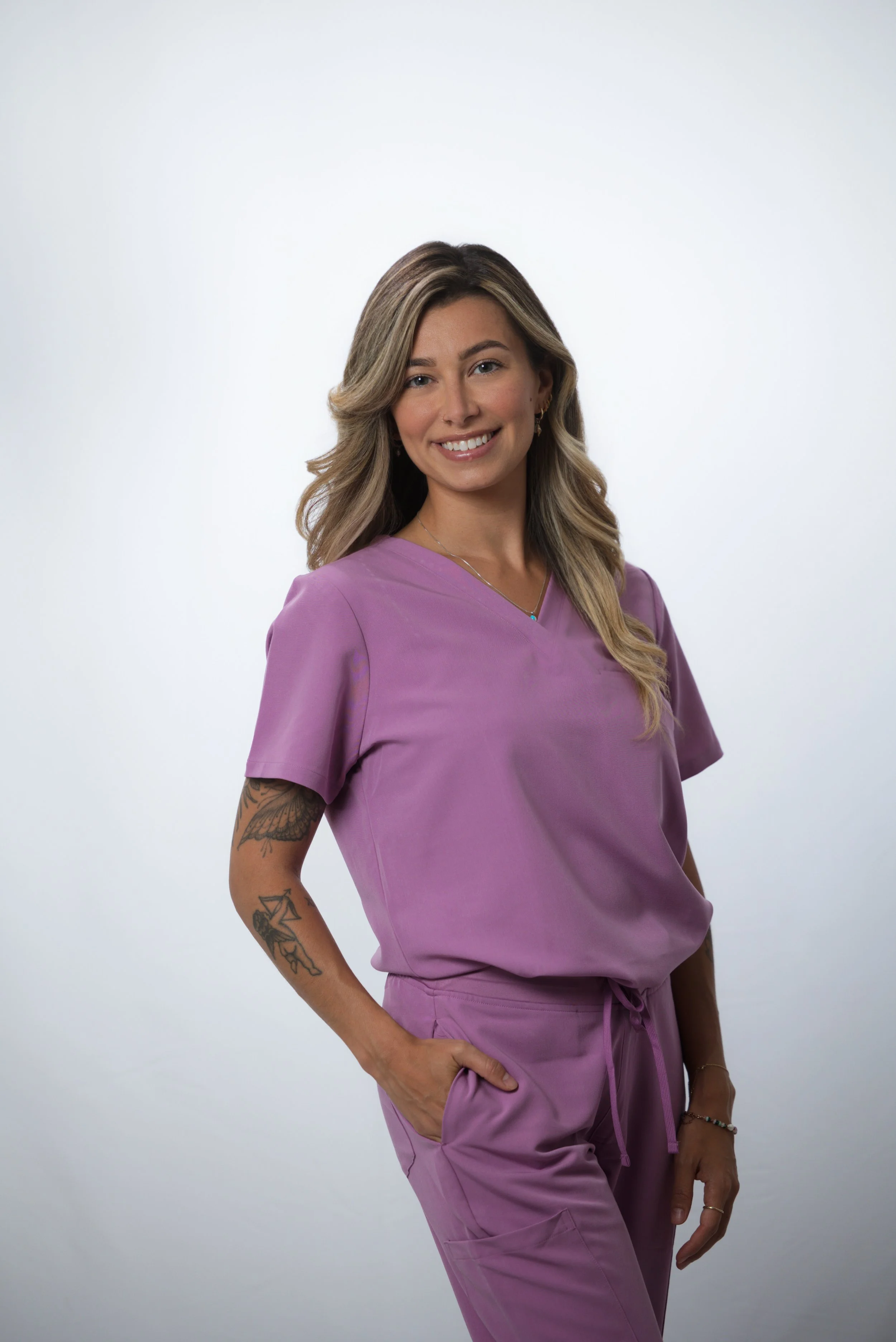 A smiling woman with blonde hair wearing purple medical scrubs, standing against a plain light background.