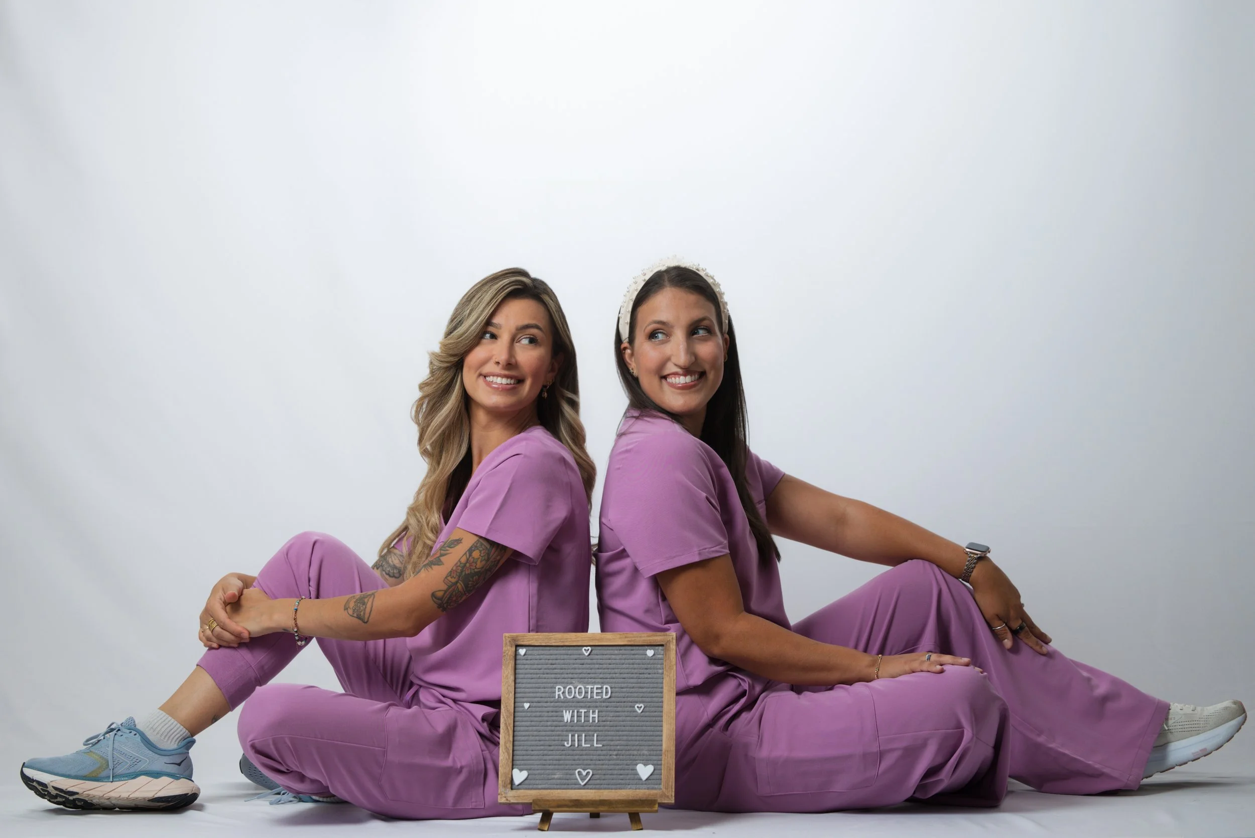 Two women wearing matching purple scrubs sit back-to-back on the floor against a plain white background, smiling and looking to the side. One has long curly hair, and the other has long straight hair with a headband. There is a small sign in front of them that reads 'Rooted with Jill' decorated with small hearts.
