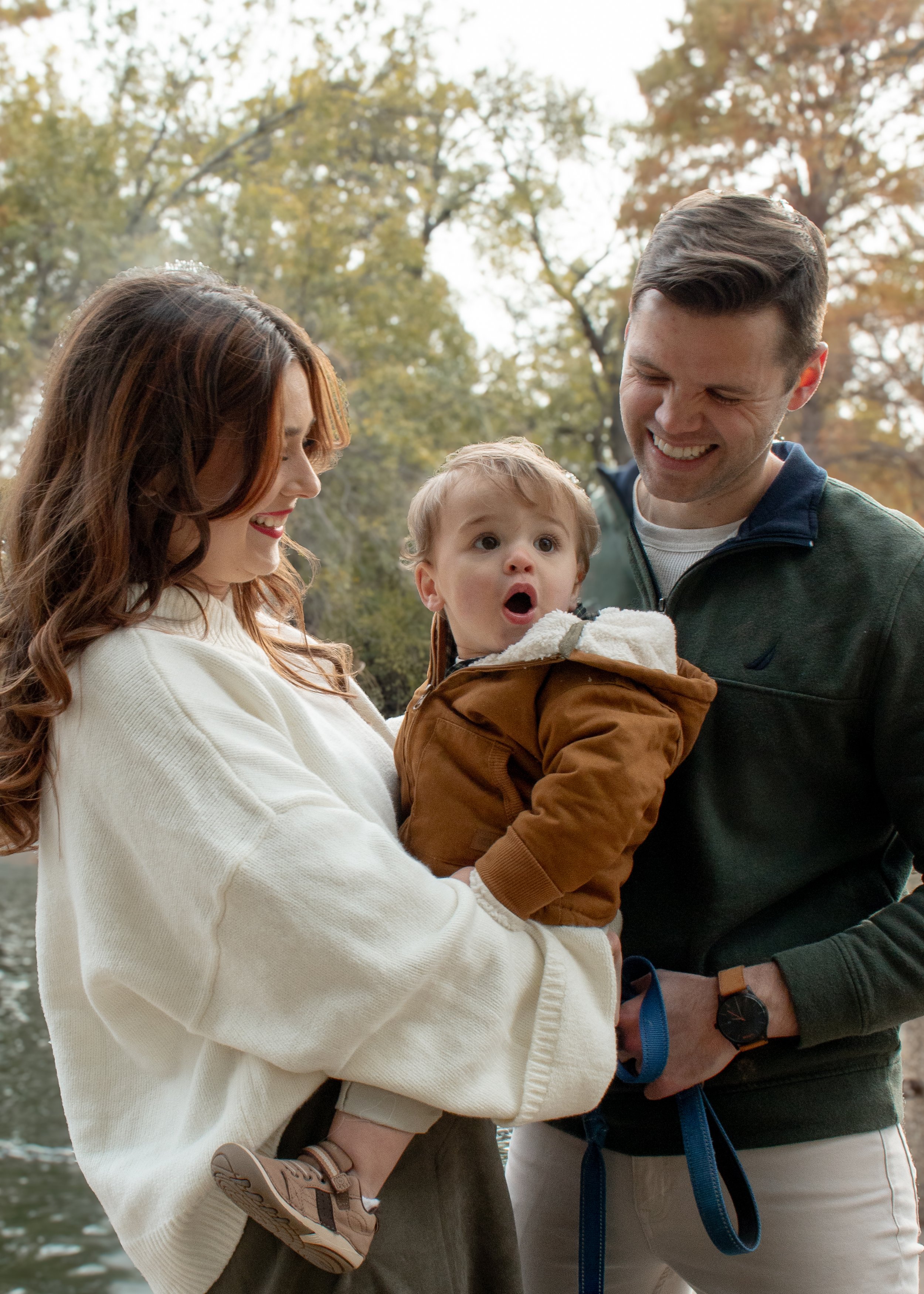 A family of three, including a mother, father, and young child, enjoying a day outdoors surrounded by autumn trees. The mother is holding the child, who is looking at the camera with an open mouth expression, while the father is smiling at the child.
