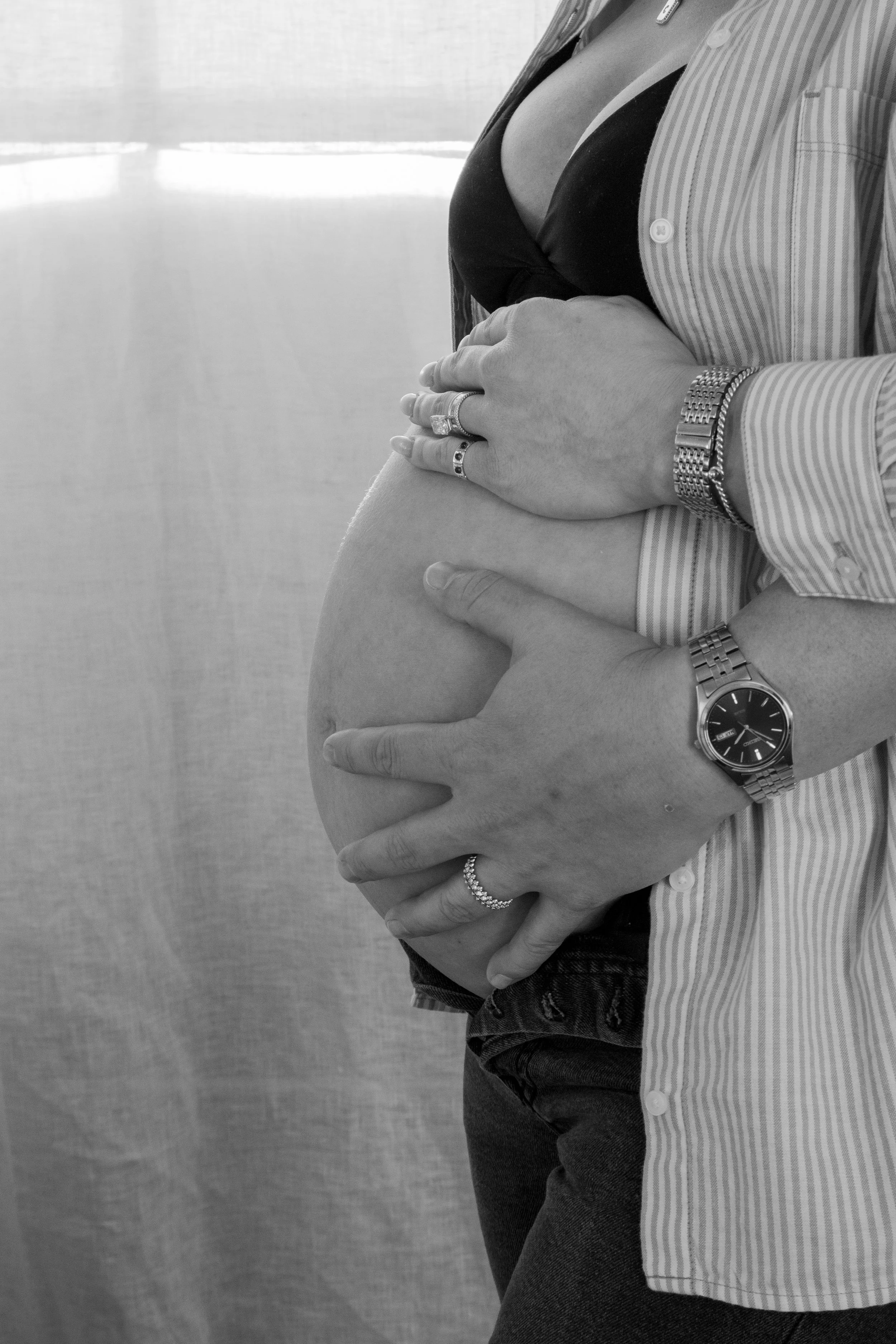 Close-up of a pregnant woman holding her belly with both hands, wearing rings and a watch, dressed in a striped shirt and jeans.