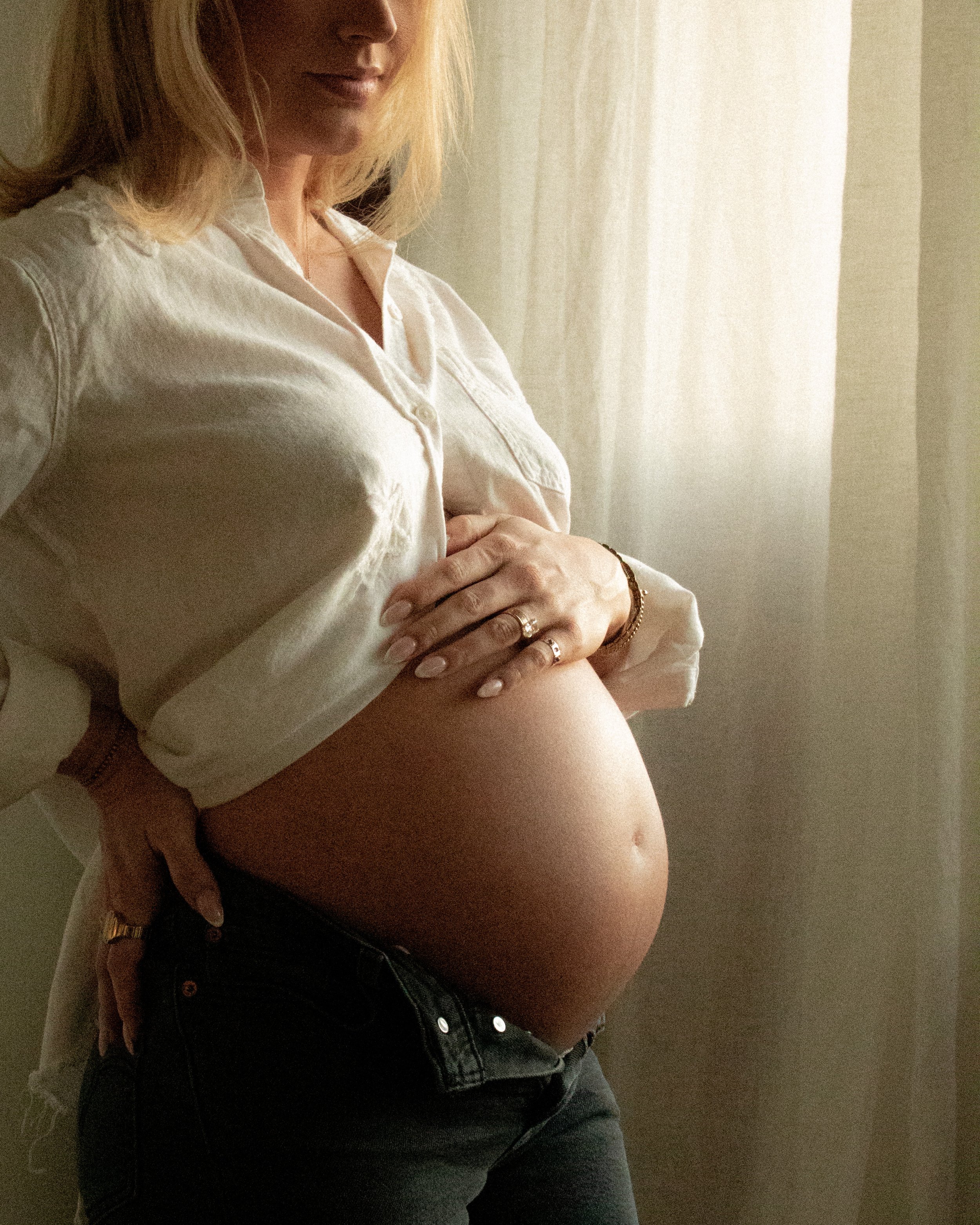 A pregnant woman in a white shirt and black jeans, standing near a window with beige curtains, with her arms crossed over her belly.