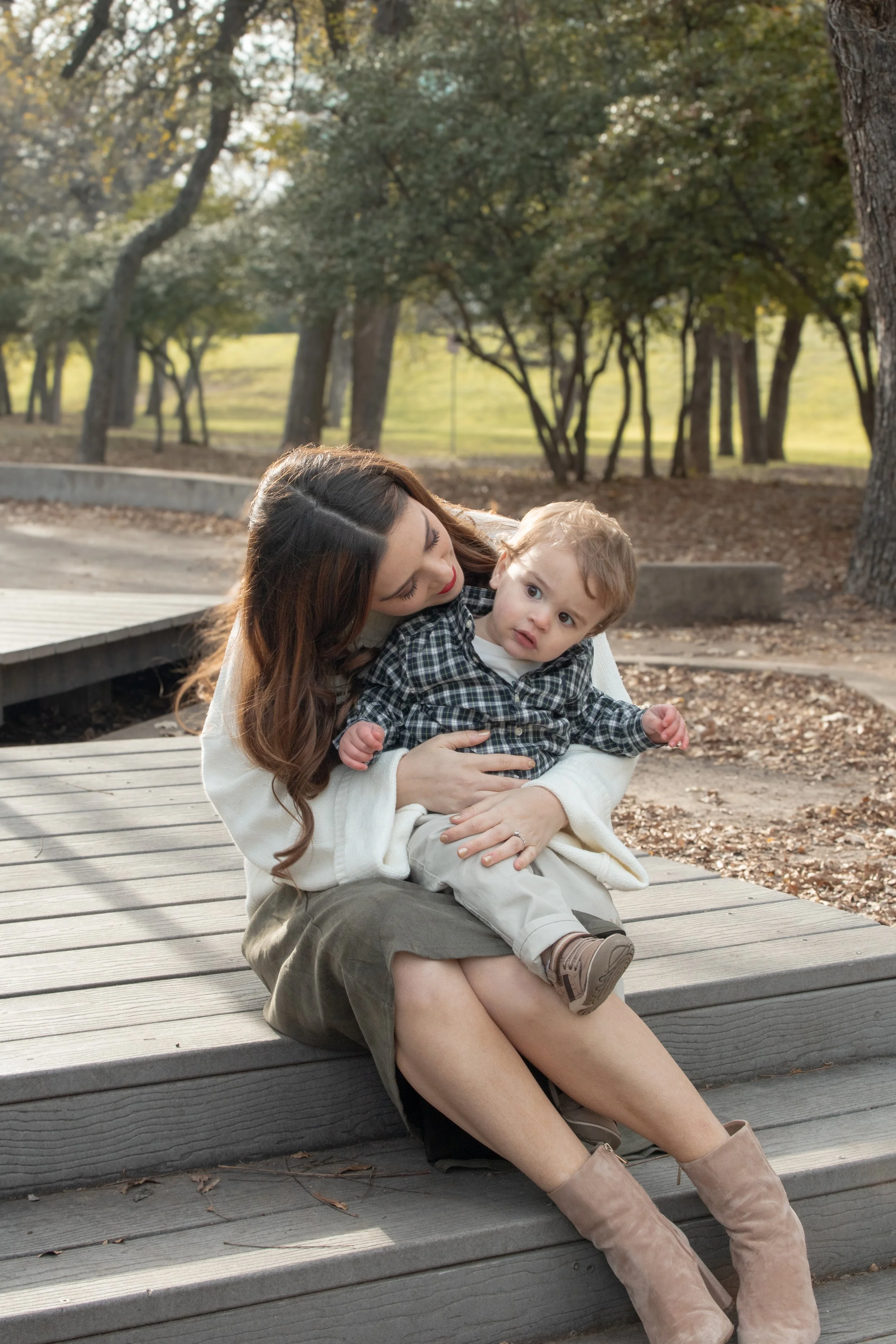 A woman with long brown hair and a light-colored sweater sitting on a wooden bench holding a young boy with blond hair and a plaid shirt at a park during autumn, with trees and fallen leaves in the background.