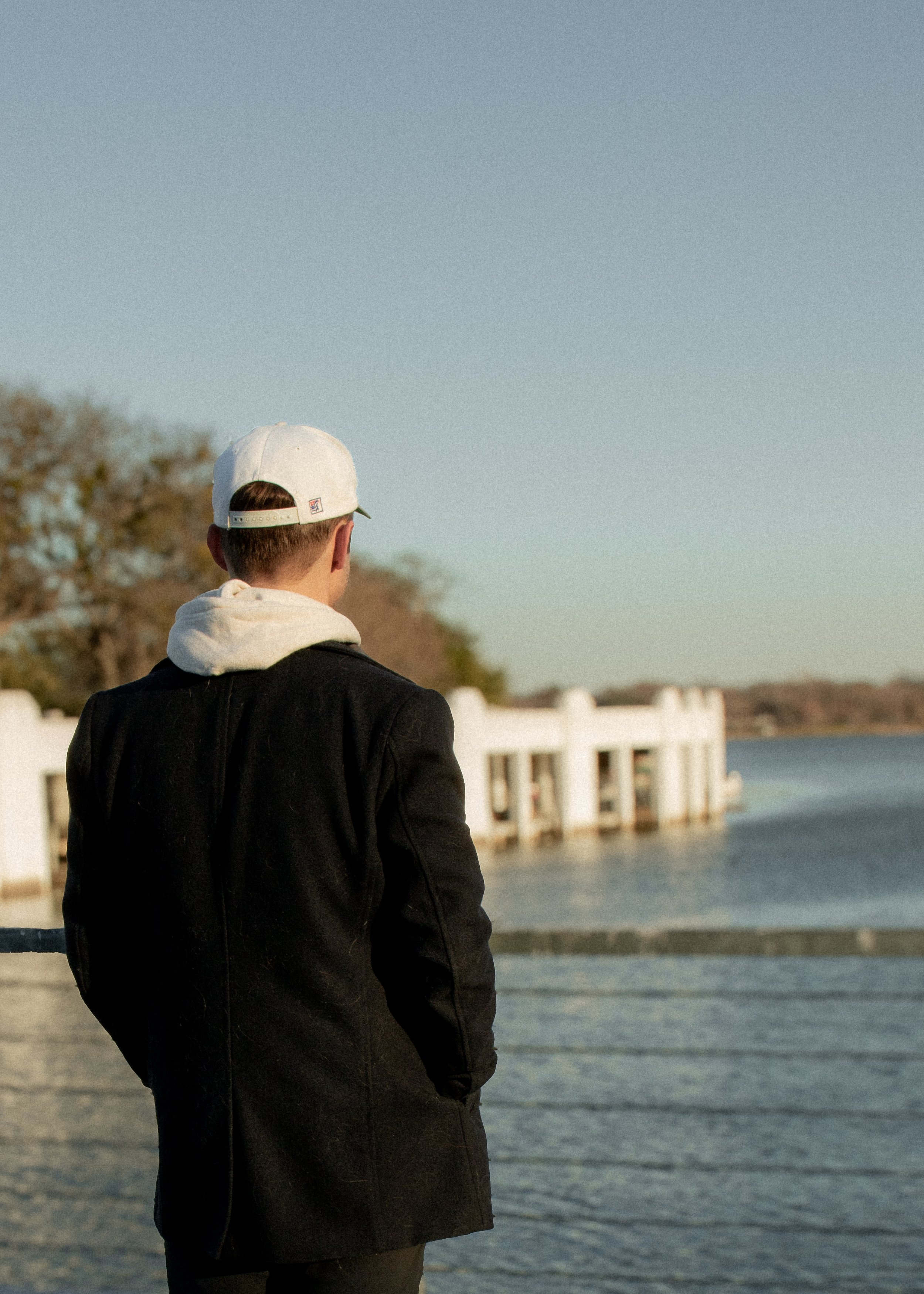 A person wearing a white cap and black jacket seen from behind, standing near a body of water with buildings in the background during dusk.