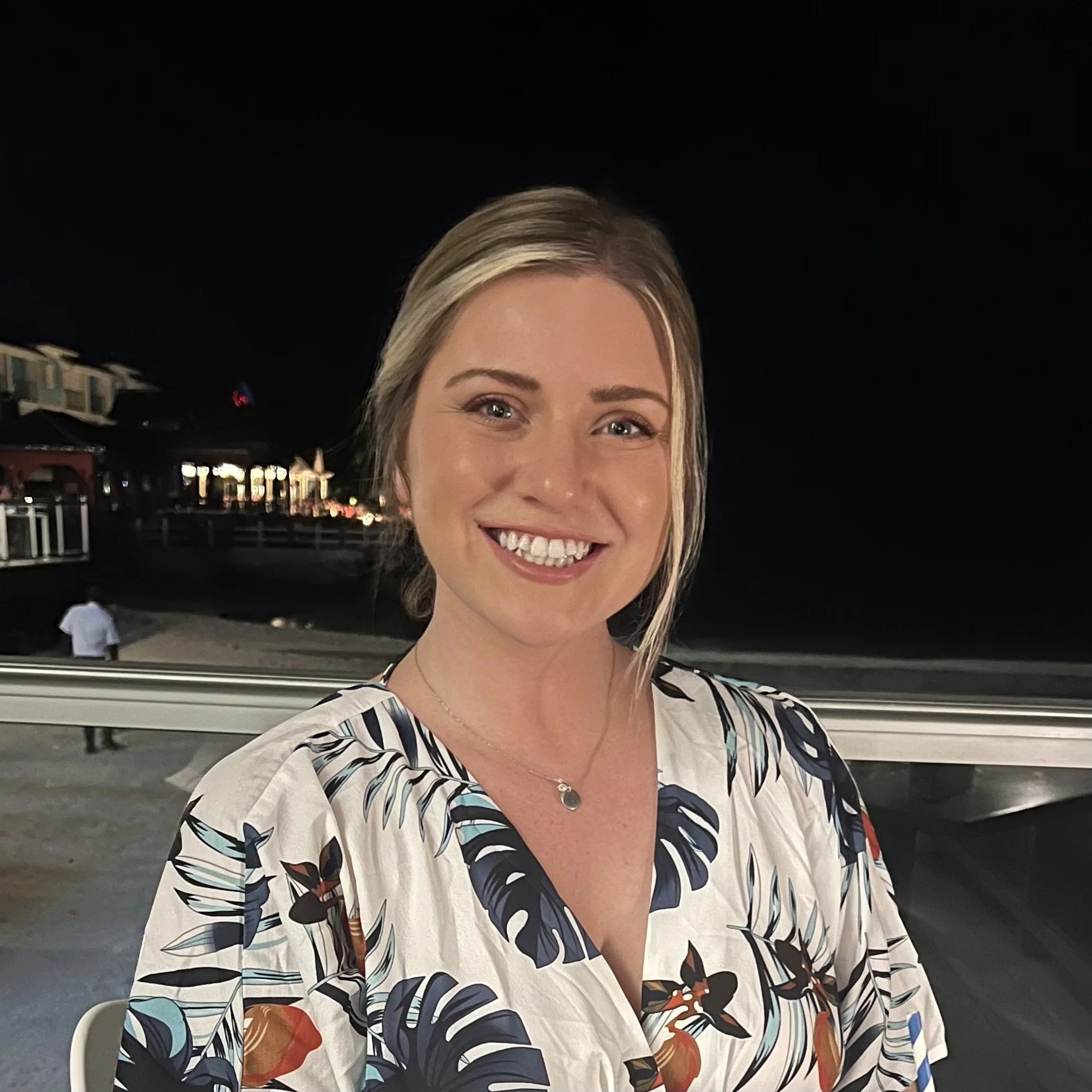 A smiling woman with blonde hair and blue eyes wearing a patterned blouse at night outdoors.