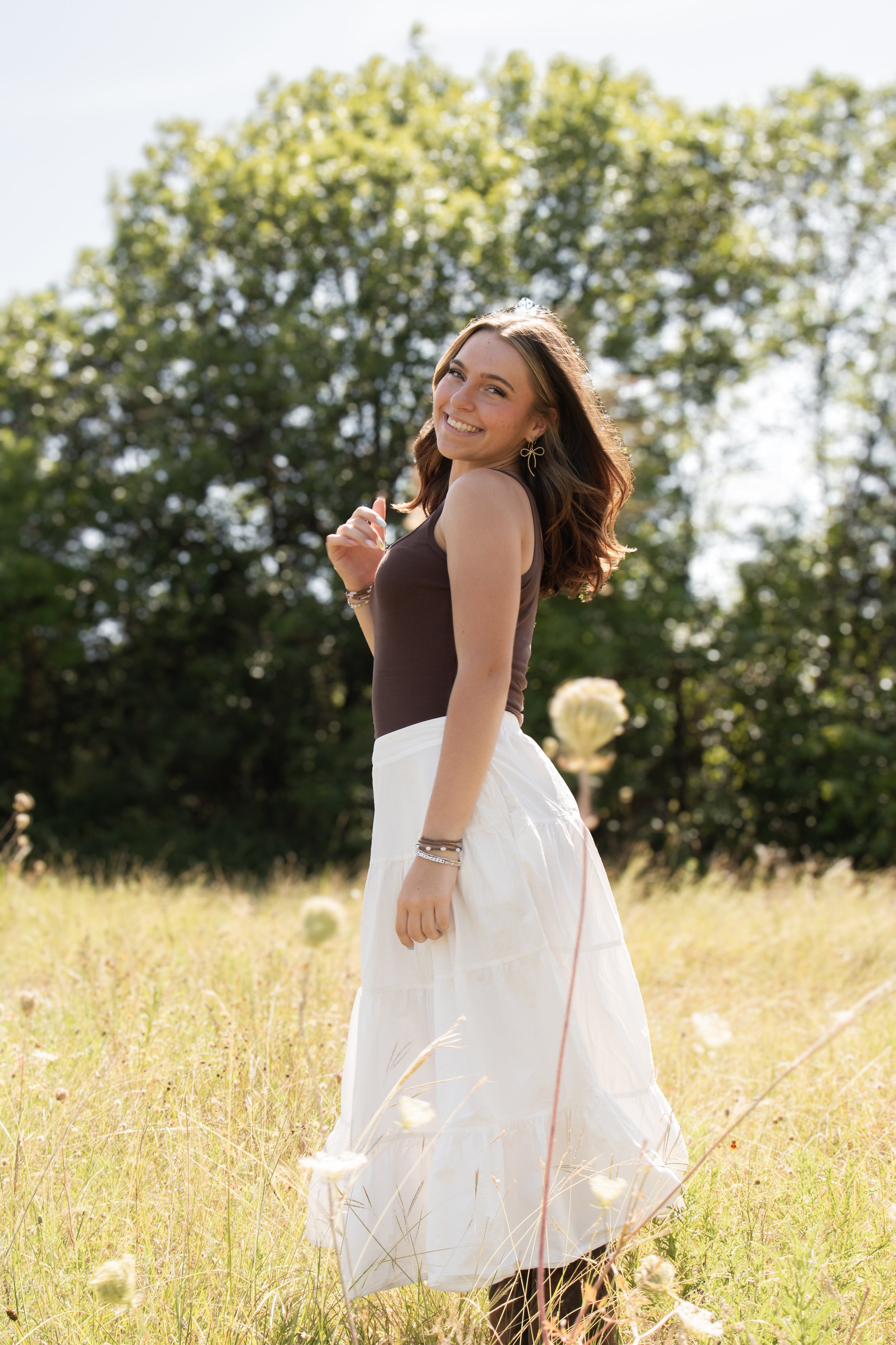 A young woman with long wavy brown hair smiling and looking back, wearing a brown sleeveless top, a white skirt, and jewelry, standing in a field of tall grass with flowers and a large tree in the background on a sunny day.