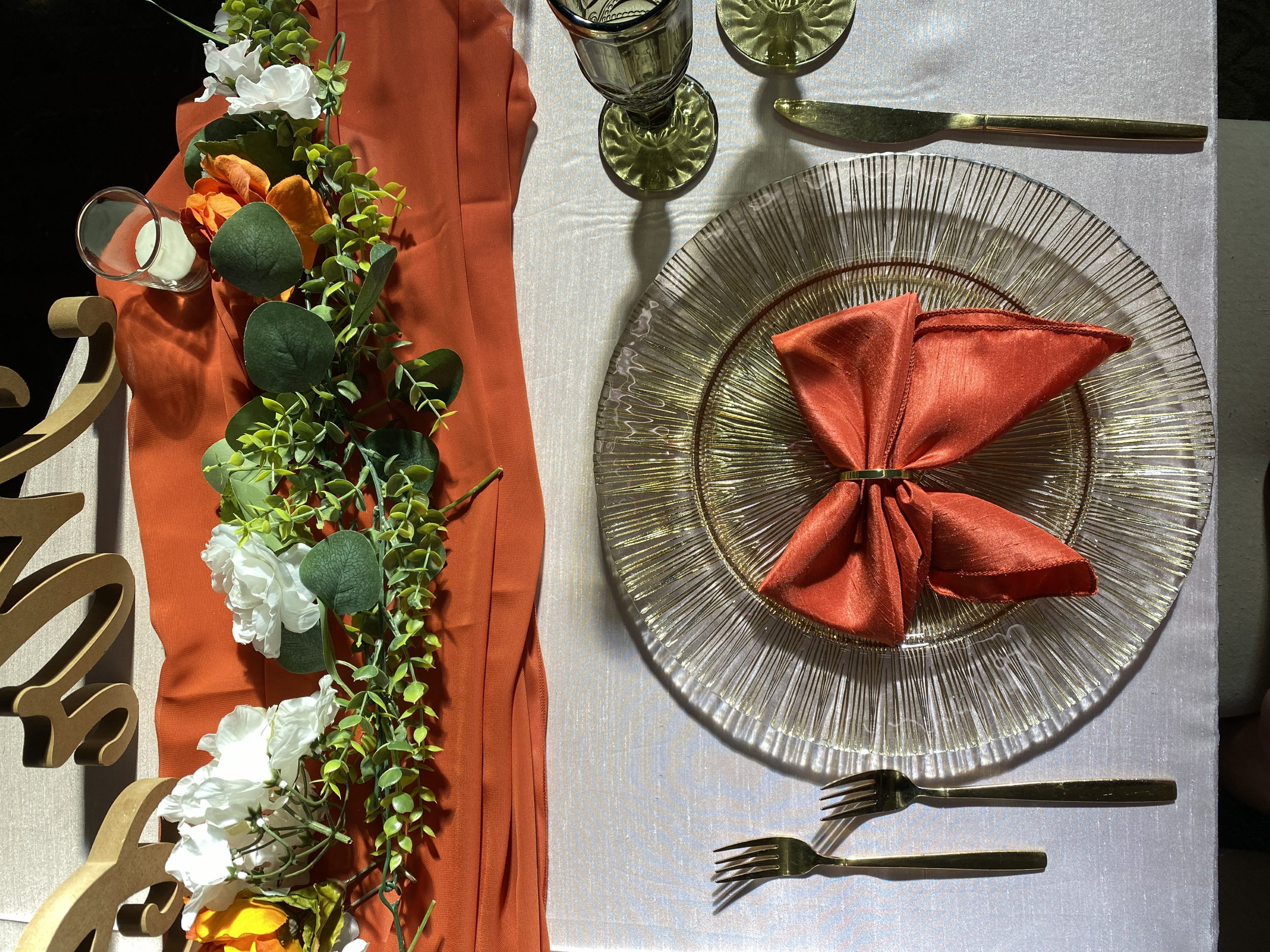 A table setting with a white tablecloth, a gold-rimmed glass plate holding a folded copper-colored cloth napkin, and two gold forks. There is a green glass with a lime slice on the rim, a green glass with a black drink, and a knife. An orange cloth runs across the table with a floral garland of white, orange, and green foliage.