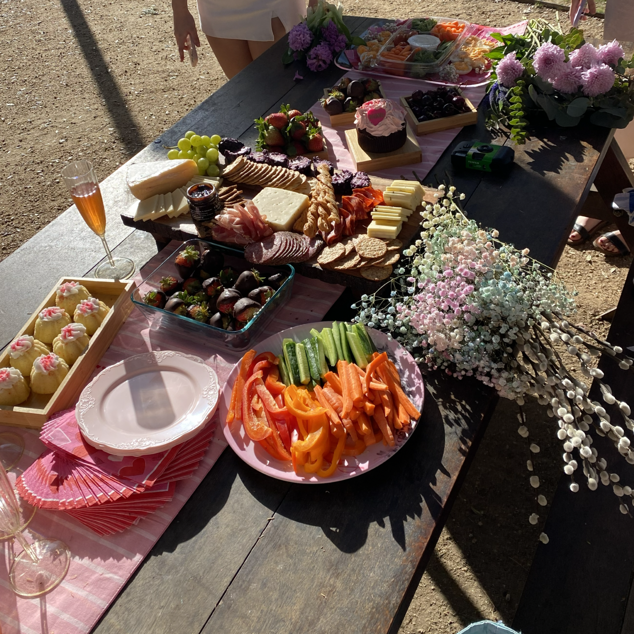 A table set outdoors with various food items including cheese, strawberries, grapes, sliced vegetables, cupcakes, and flowers, with people in the background.
