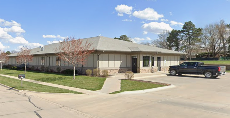 A single-story beige building with a gray roof, surrounded by grass and trees, with a parking lot and a black pickup truck in front.