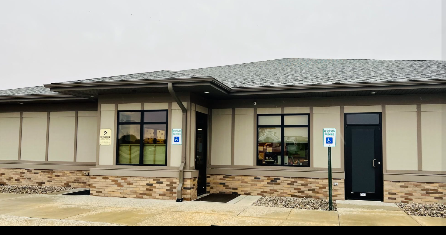 Front view of a beige commercial building with two large windows and a black door, featuring two space markers for accessible parking, a "reserved parking" sign, and a "no parking" sign, with a wet sidewalk and gravel area in front.