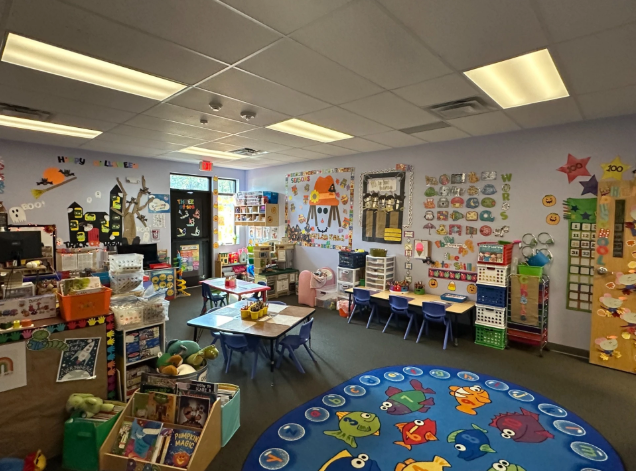 A colorful classroom with alphabet-themed wall decorations, tables, chairs, and toys, including a large circular rug with animal numerals designed for young children.