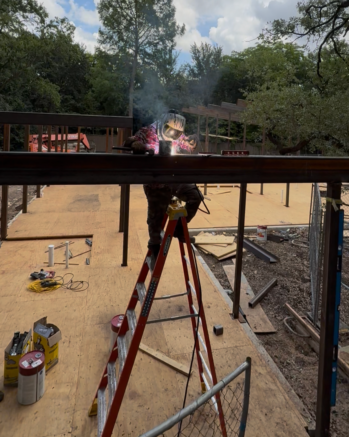 A construction worker welding metal at a building site on a wooden platform outdoors, with tools and materials scattered around.