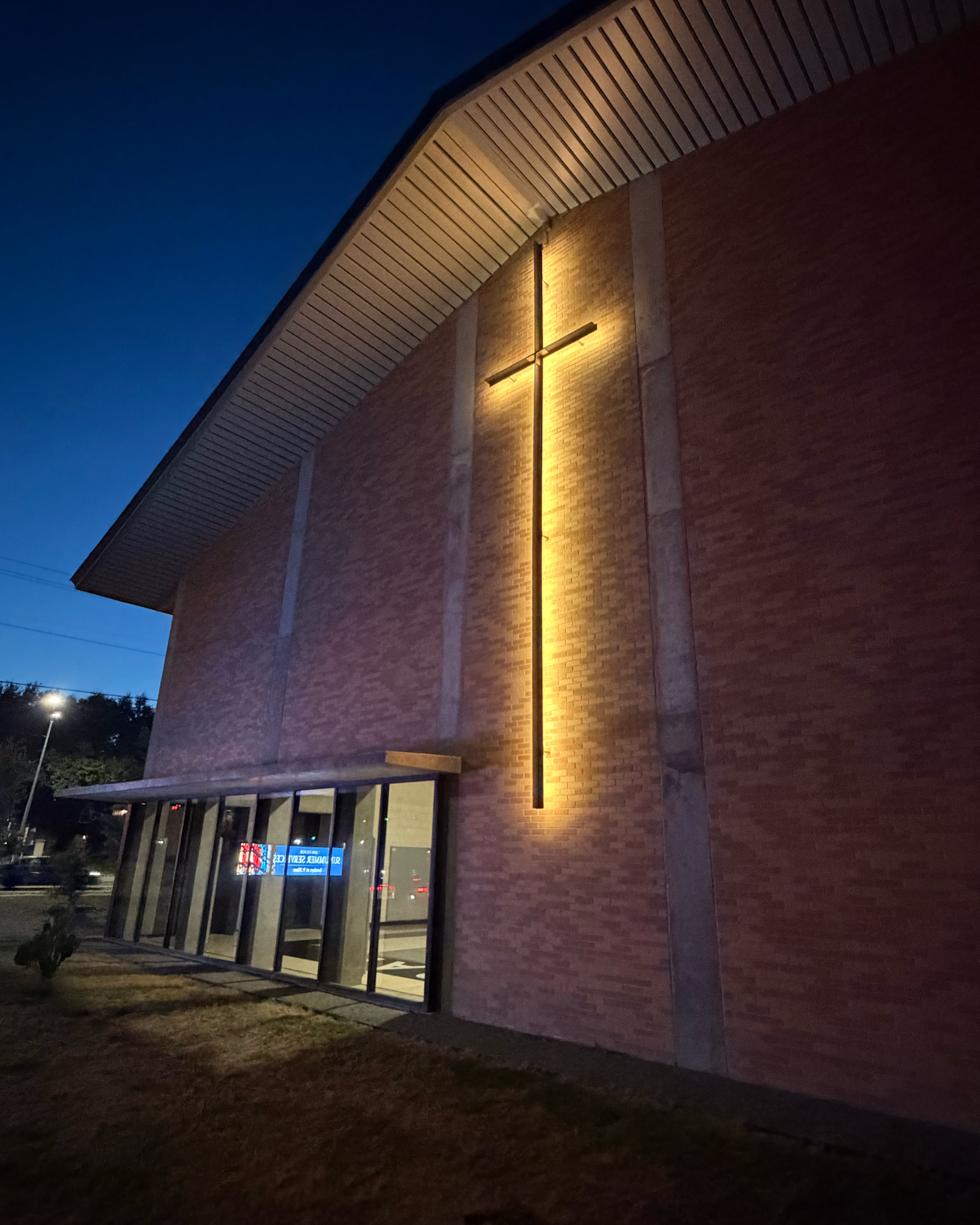 Exterior of a church building at dusk with a large illuminated custom steel cross on the brick wall and a glass entrance door part of the building.