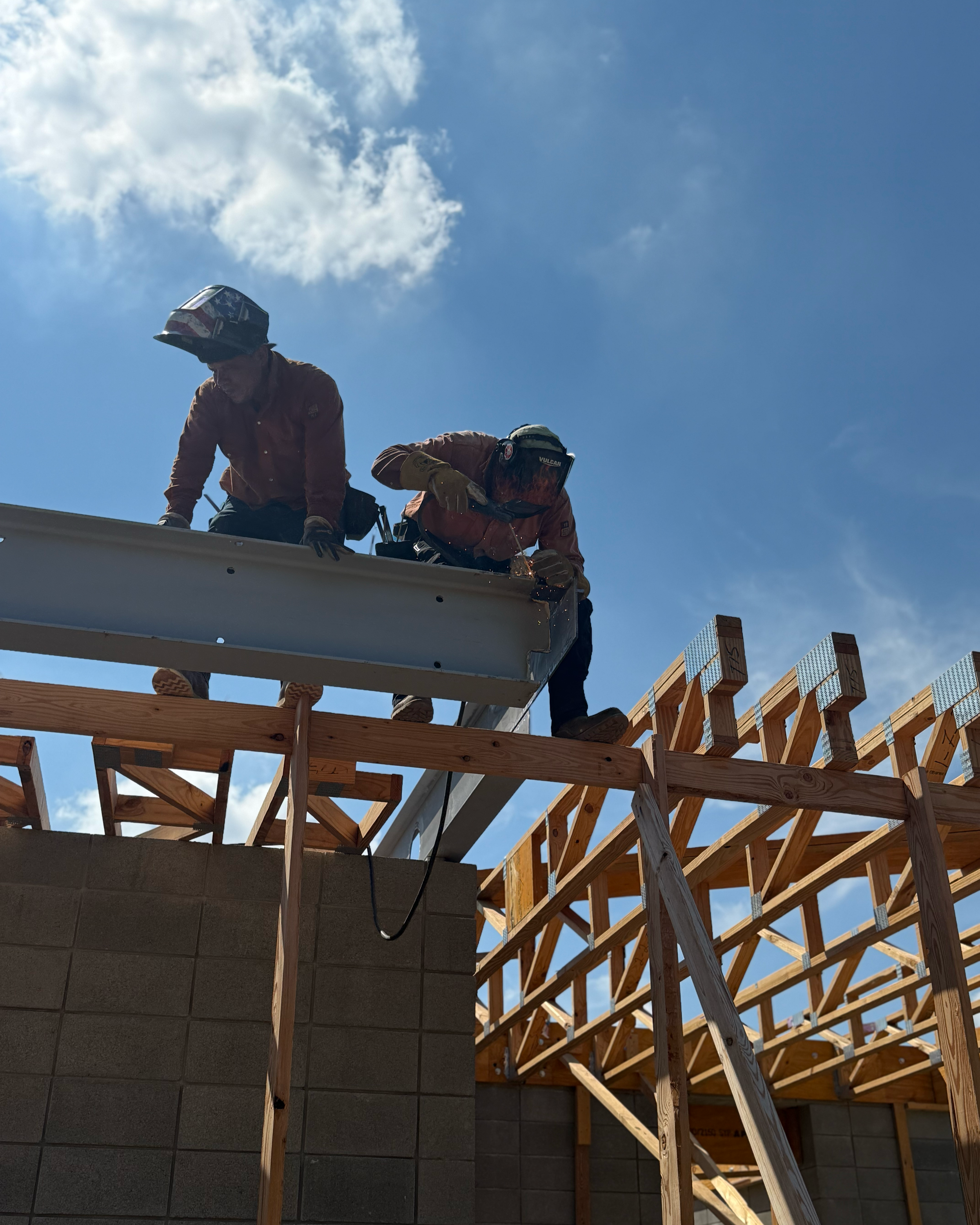 Construction workers on a building site, working on a wooden roof framework with a blue sky and clouds in the background.