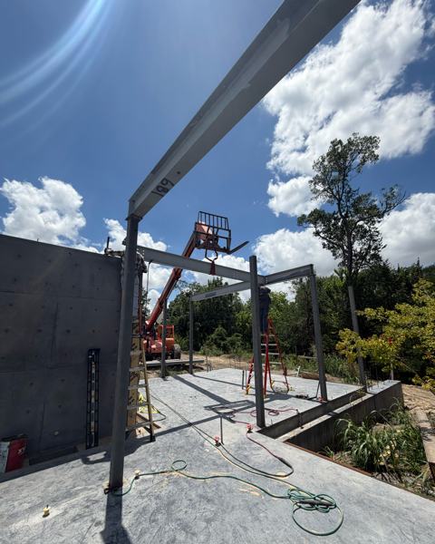 Construction site with steel framing, a cherry picker, and a partially built structure under a blue sky with clouds.