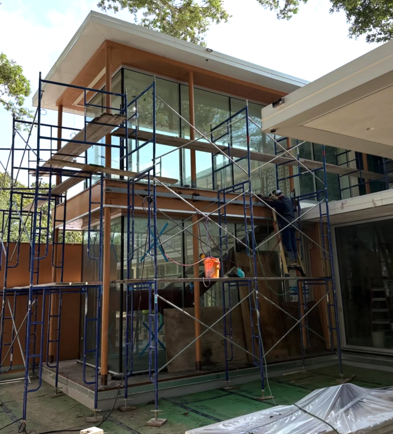 Construction workers installing structural steel on a modern home with large glass windows, wood accents, and scaffolding.