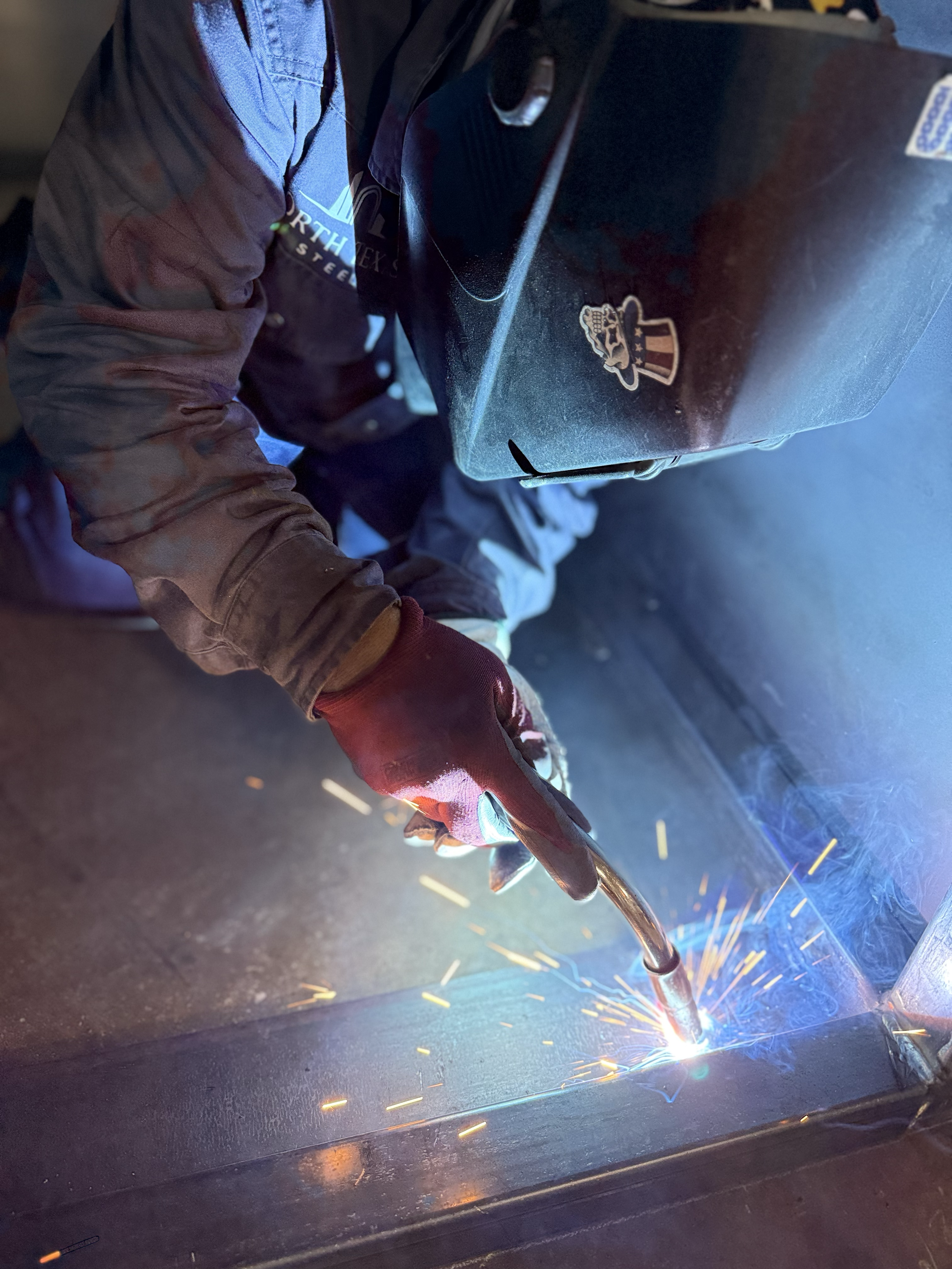 A person welding steel, wearing a brown jacket, gloves, and a welding helmet, with sparks flying as they work on a metal piece.