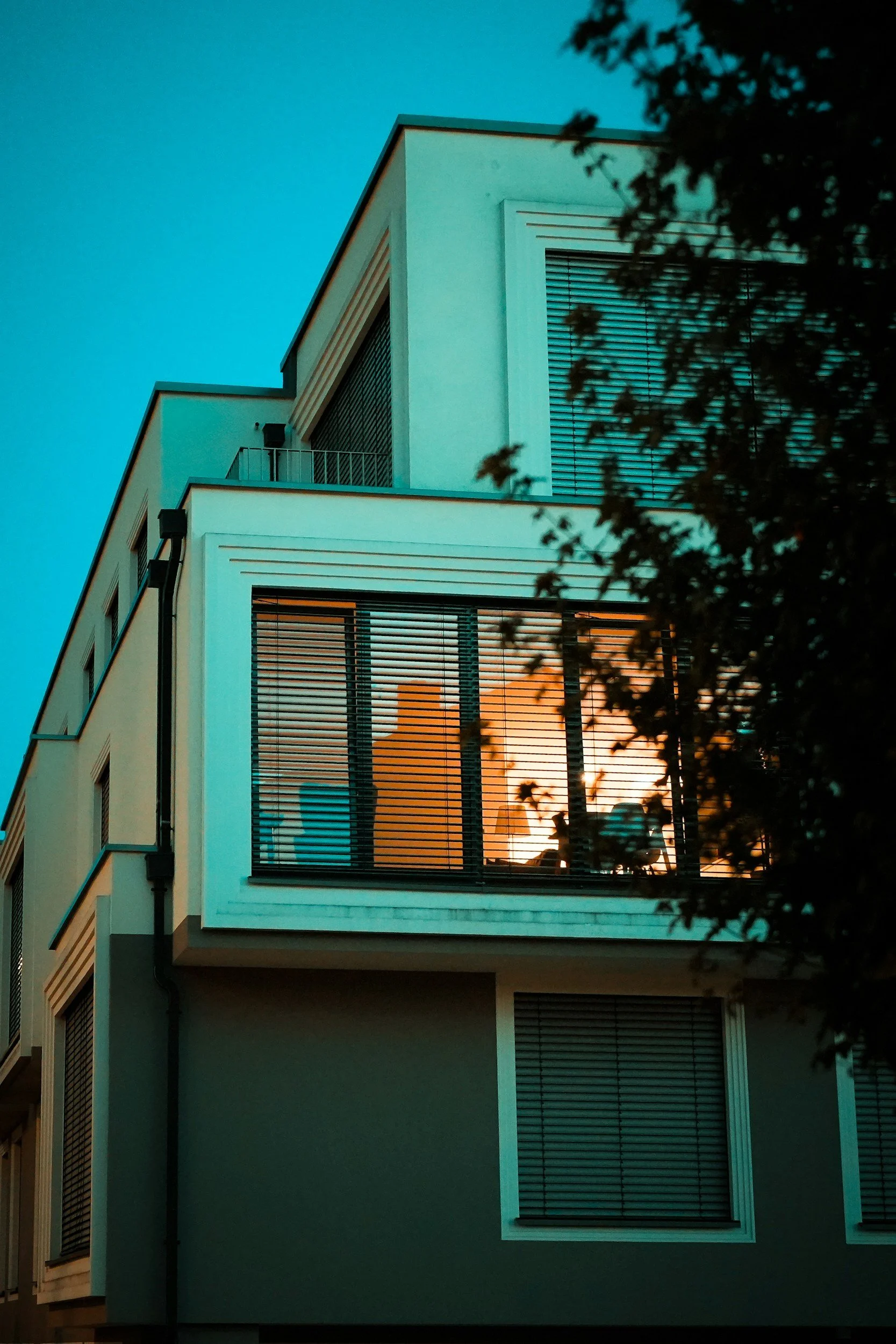 Modern multi-story building at sunset with illuminated woman sitting on balcony. Dark tree branches in foreground.