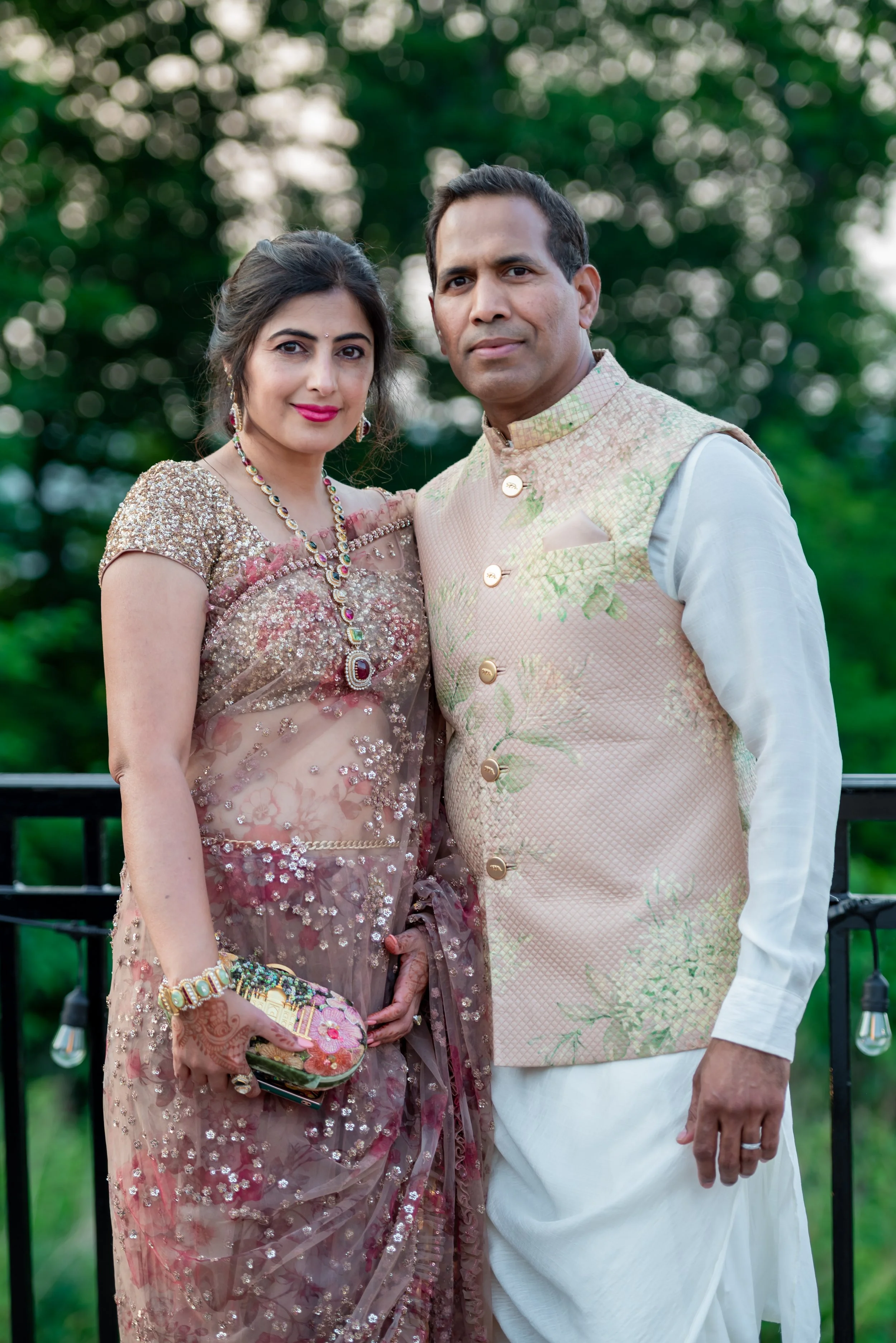 A couple dressed in traditional Indian attire, standing outdoors with greenery in the background. The woman is wearing a embellished pink saree with jewelry, and the man is wearing a beige embroidered vest over a white kurta.