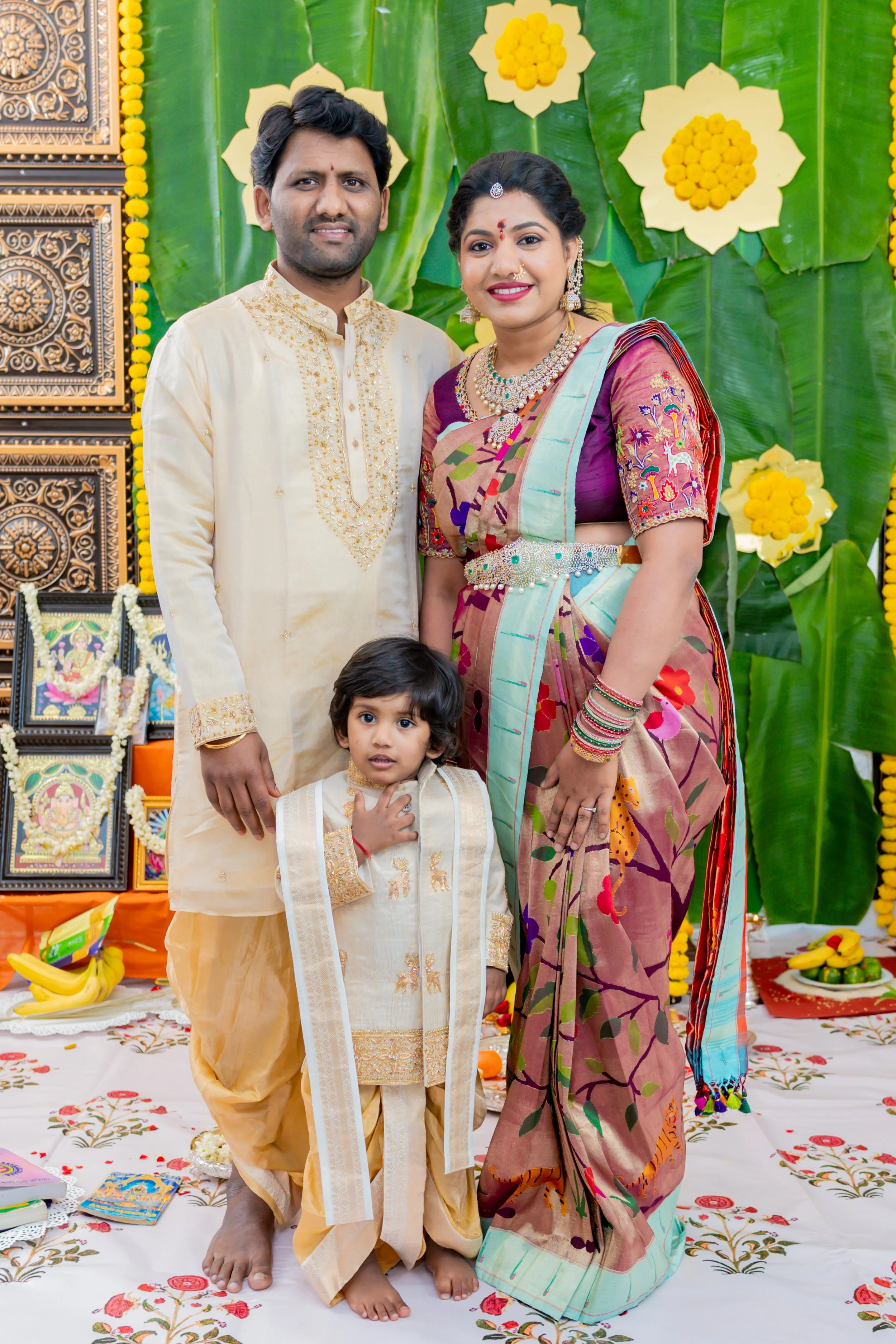 A family at a traditional Hindu ceremony, with a man, woman, and child dressed in colorful traditional Indian attire, standing in front of a decorated backdrop with green leaves, yellow flowers, and religious images.