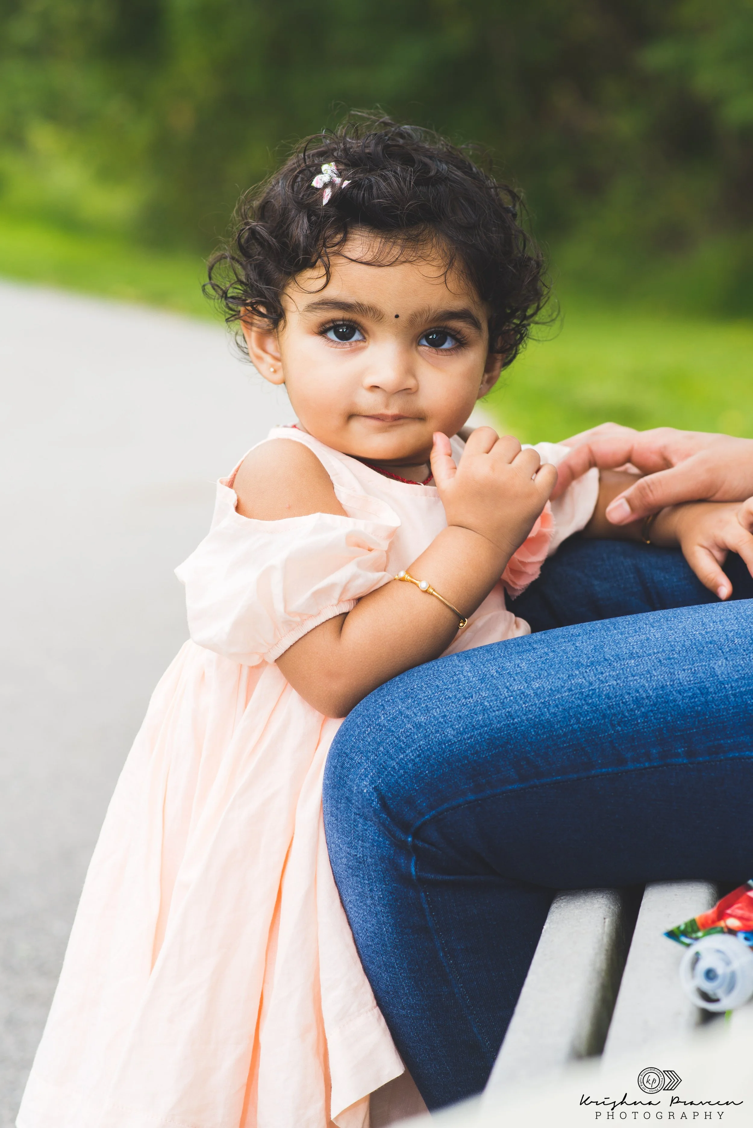 Little girl with curly hair, blue eyes, and a small black bindi on her forehead, wearing a peach-colored dress, holding hands with an adult sitting on a park bench.