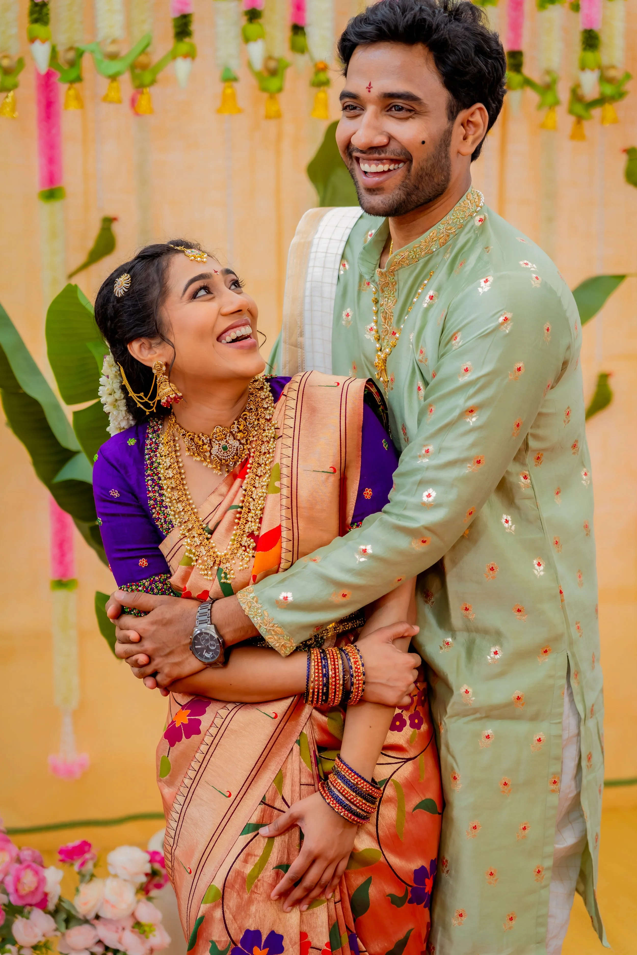 A joyful Indian couple celebrating at a traditional event, dressed in colorful traditional attire. The woman wears a saree with floral patterns and is adorned with gold jewelry and flowers in her hair. The man wears a light green kurta with red and g