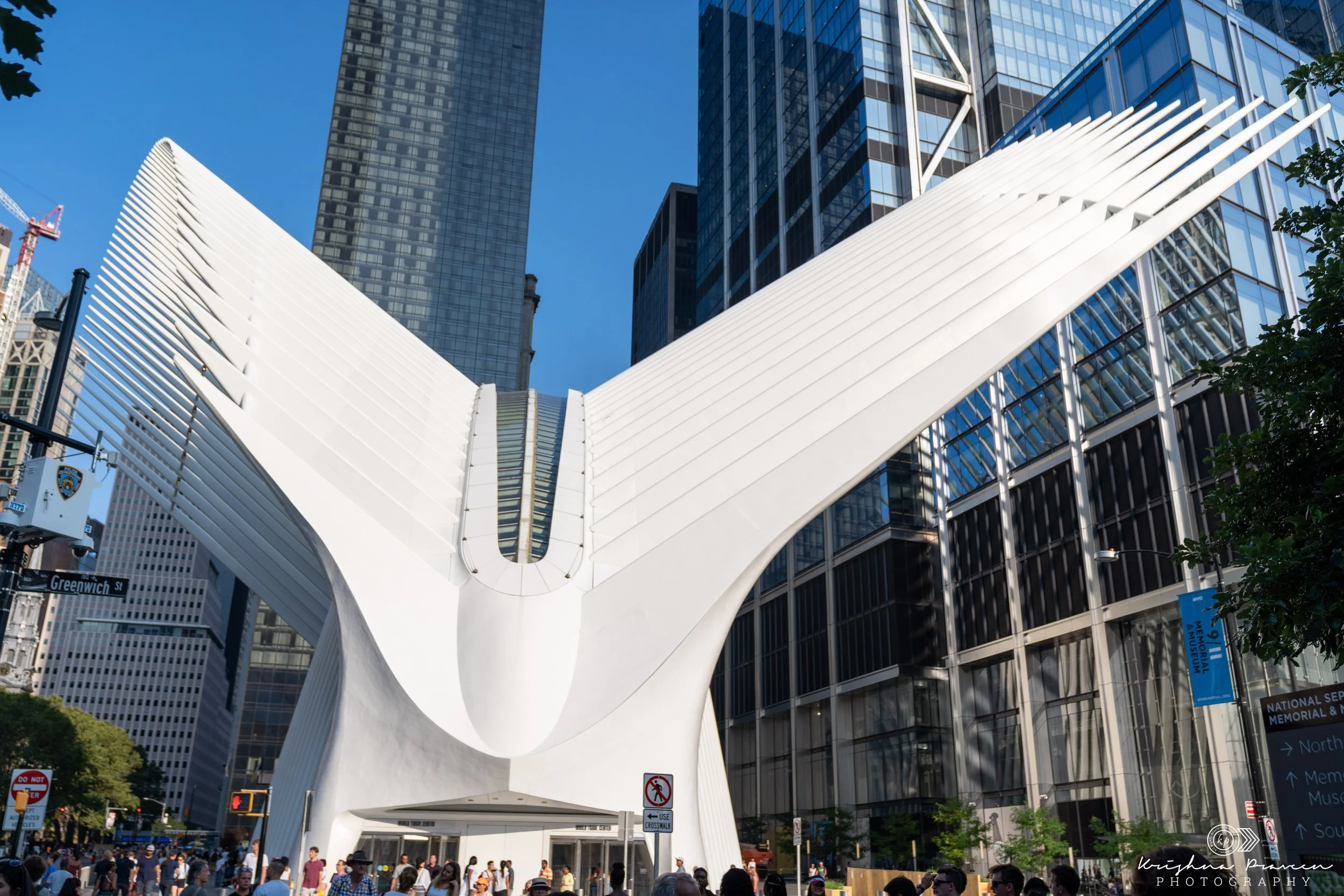 The image shows a large white modern architectural structure with steep, sloped sides, situated in an urban area with tall office buildings. There are people walking underneath and around the structure, and trees are visible on the sides. A street si