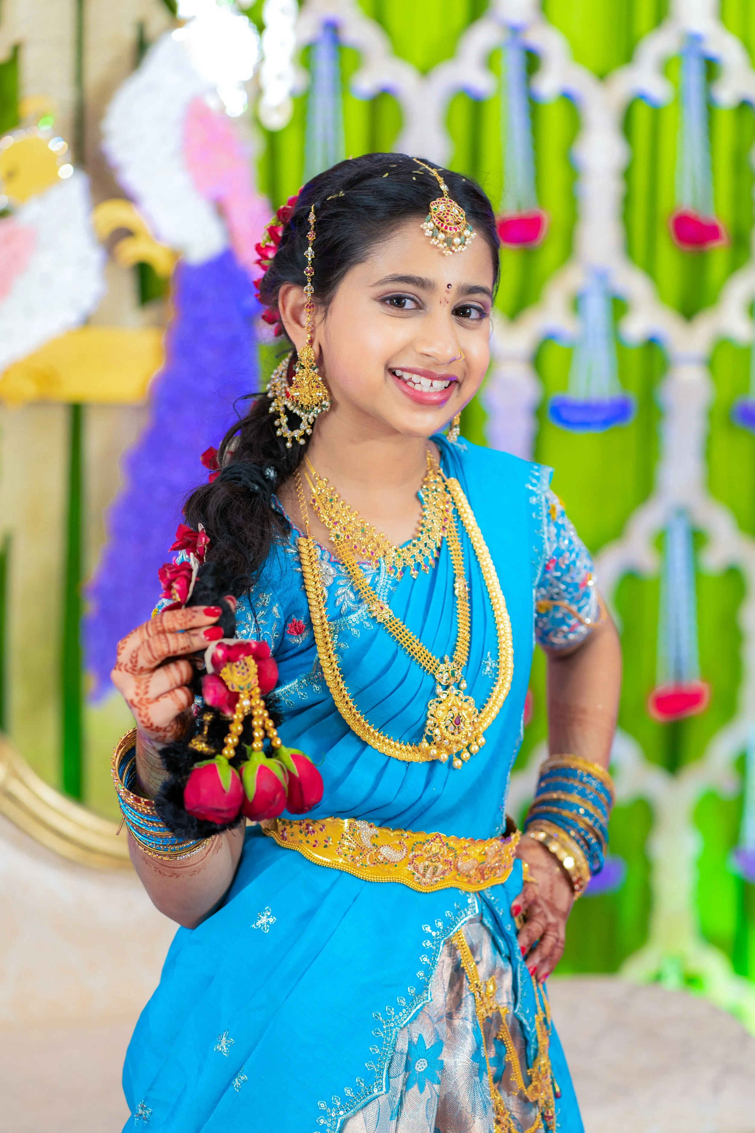 A young woman dressed in traditional blue Indian attire with gold jewelry, smiling and holding pink rose hair accessories, standing in front of colorful floral decorations.