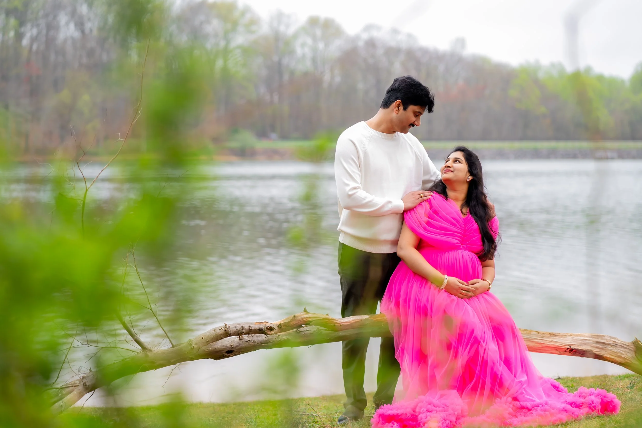 A couple stands by a river, the woman in a pink flowing dress and the man in a white sweater, gazing at each other with a smiling expression.