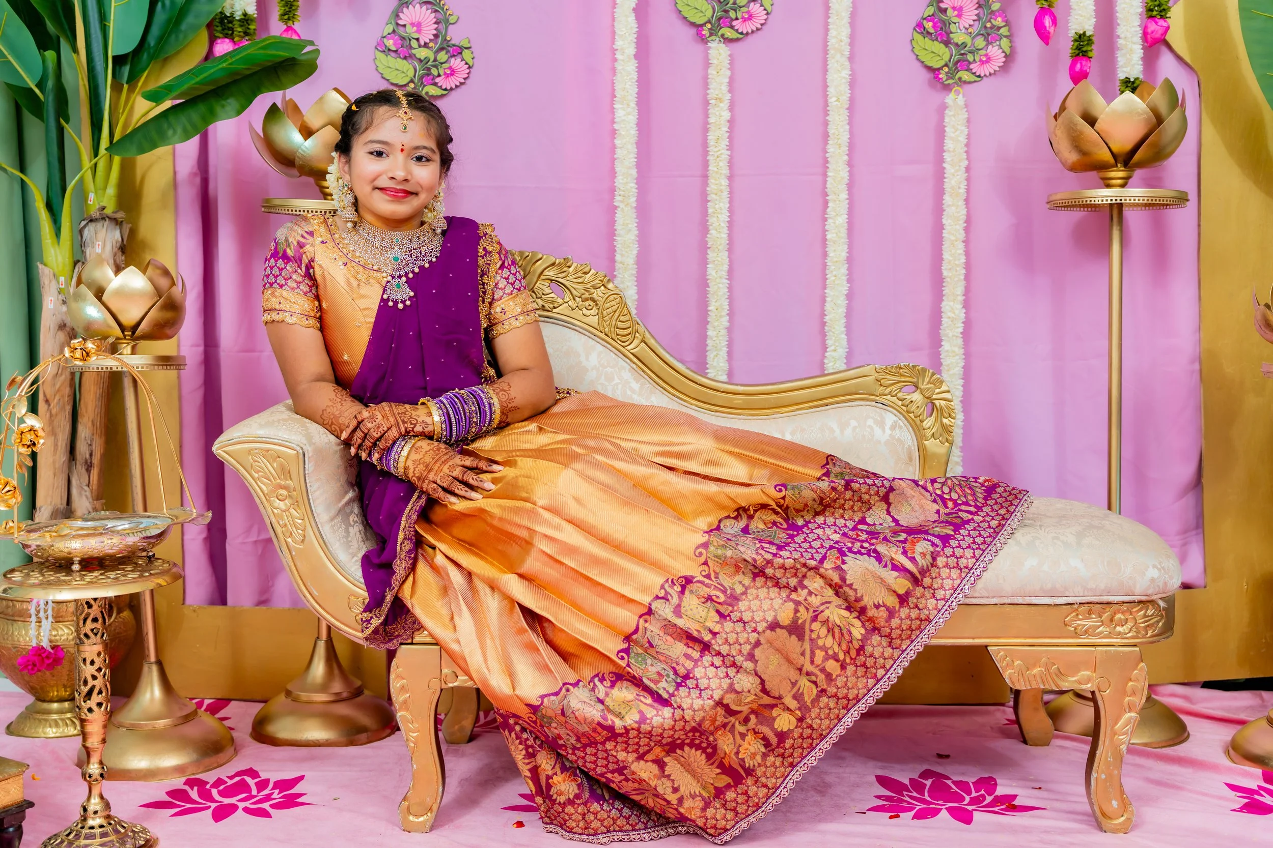 A young girl dressed in traditional Indian attire, sitting on a gold ornate sofa amid pink and yellow decorations with floral and leaf accents, gold lotus-shaped decor, and pink flower petals on the floor.