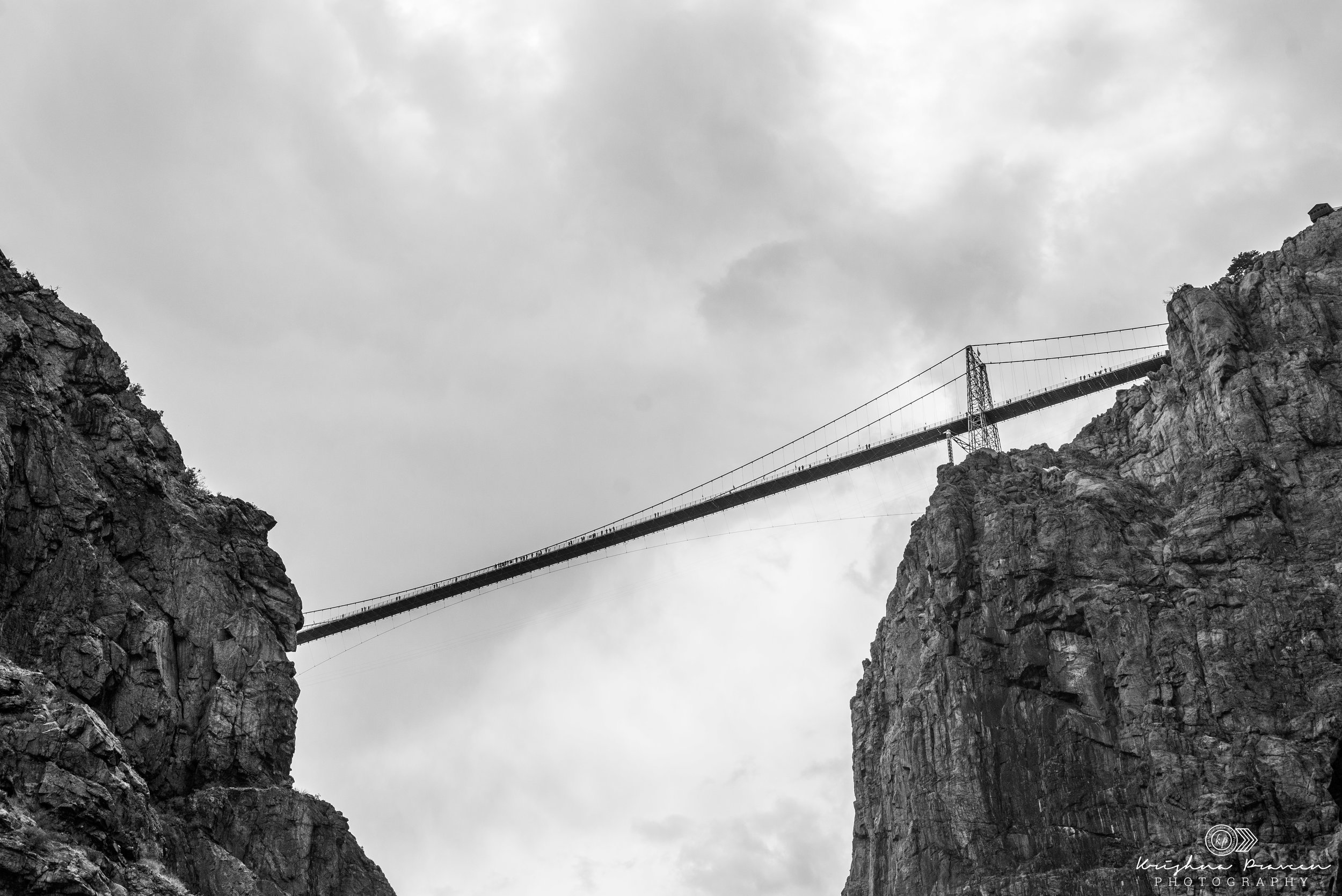 A black and white photograph of the Royal Gorge Bridge spanning between two tall rocky cliffs with a cloudy sky above.