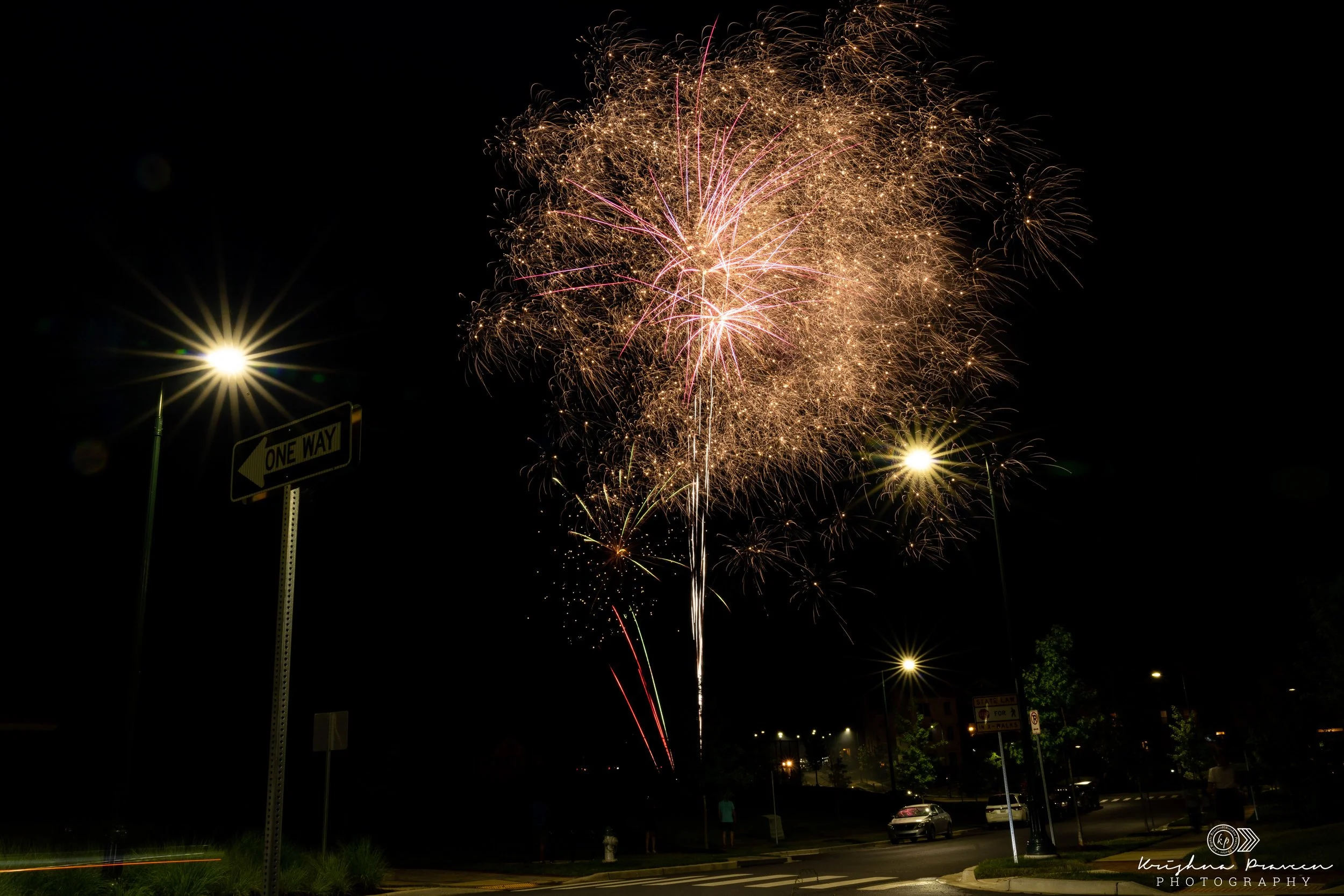 Fireworks exploding in the night sky over a parking lot with people and cars visible below, streetlights illuminating the area.
