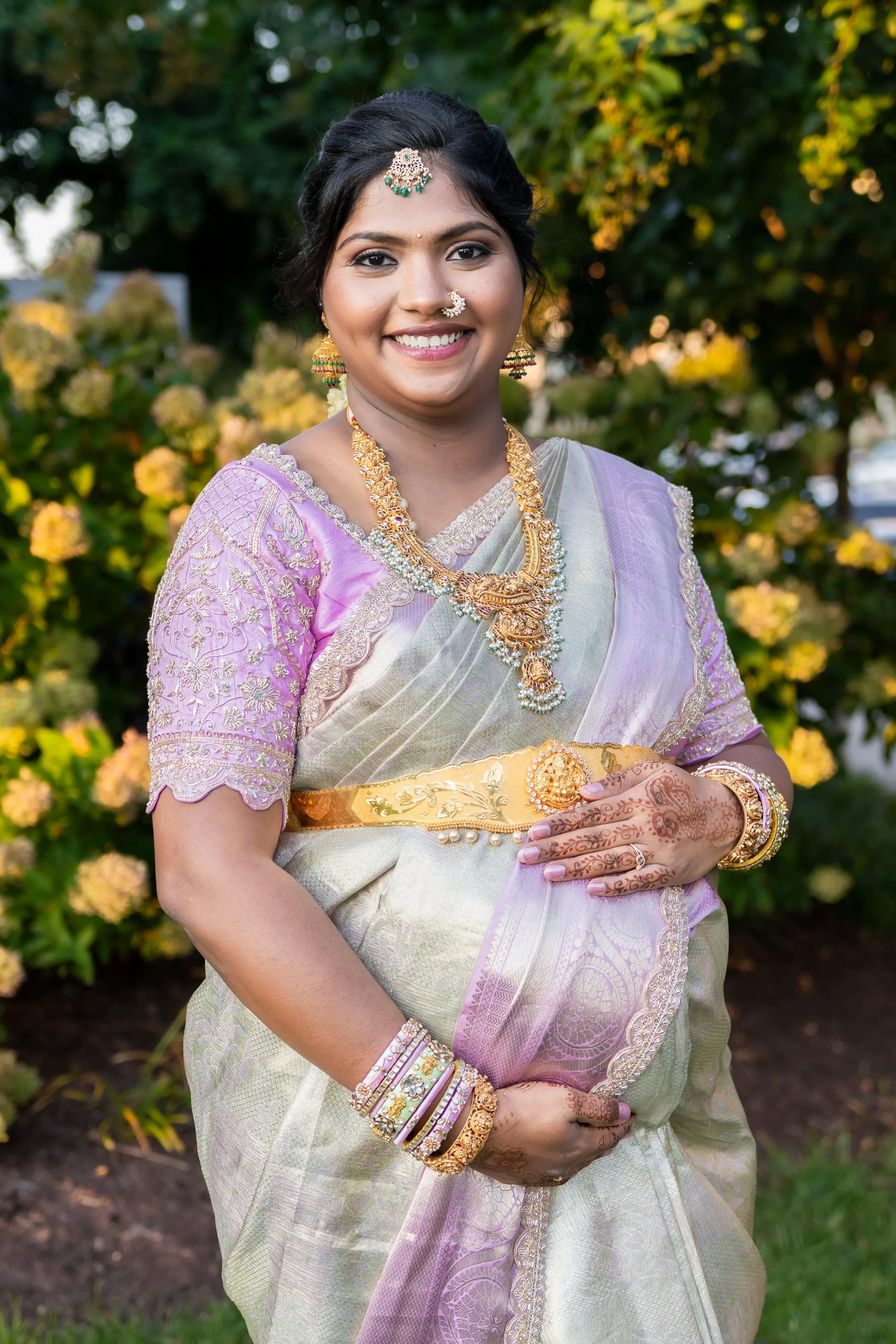 A woman dressed in a light purple and cream traditional Indian saree, adorned with gold and jewelry, smiling outdoors with greenery in the background.