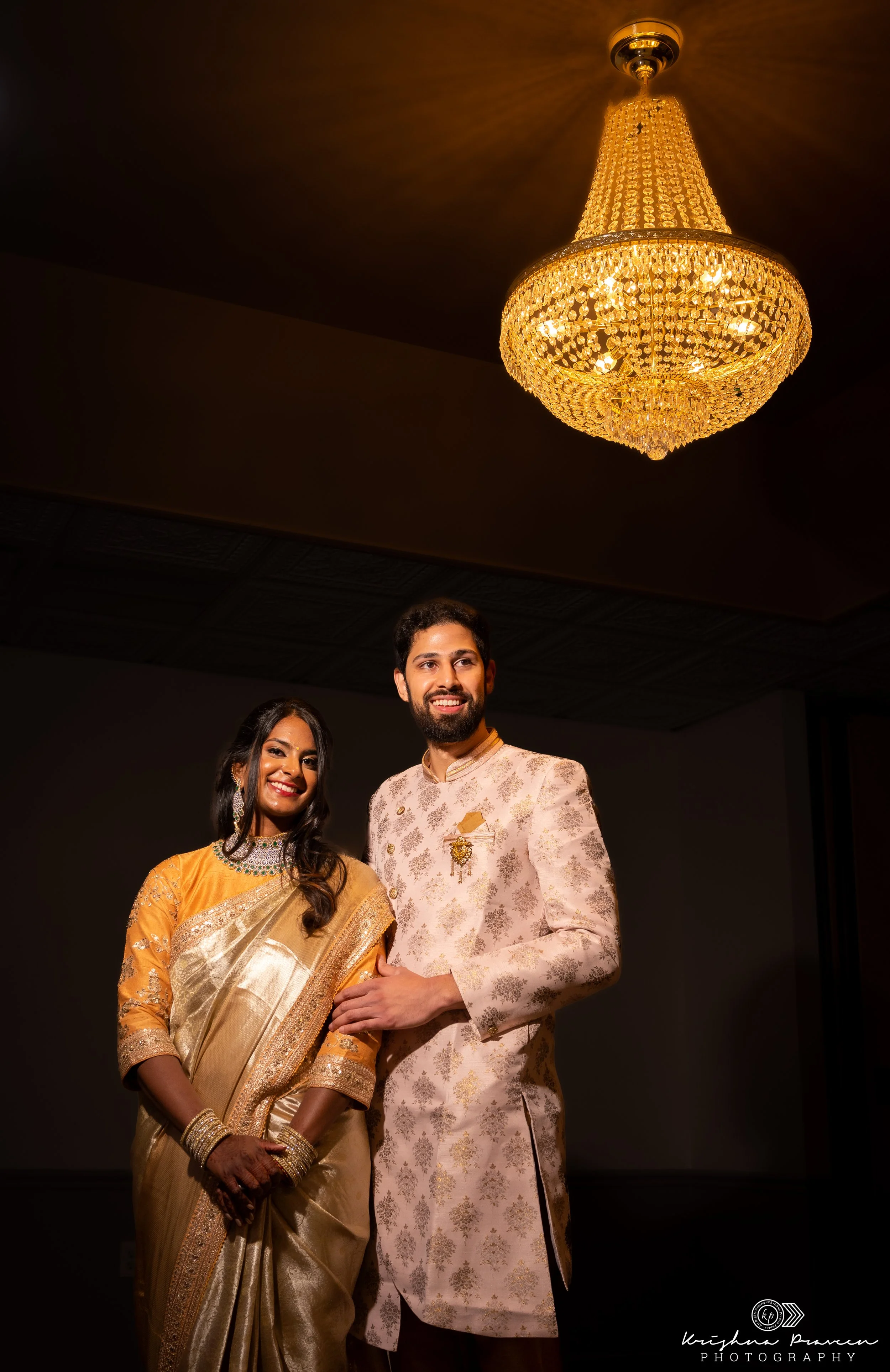 A man and woman dressed in traditional Indian attire smiling at a photographer, standing underneath a large, ornate chandelier.