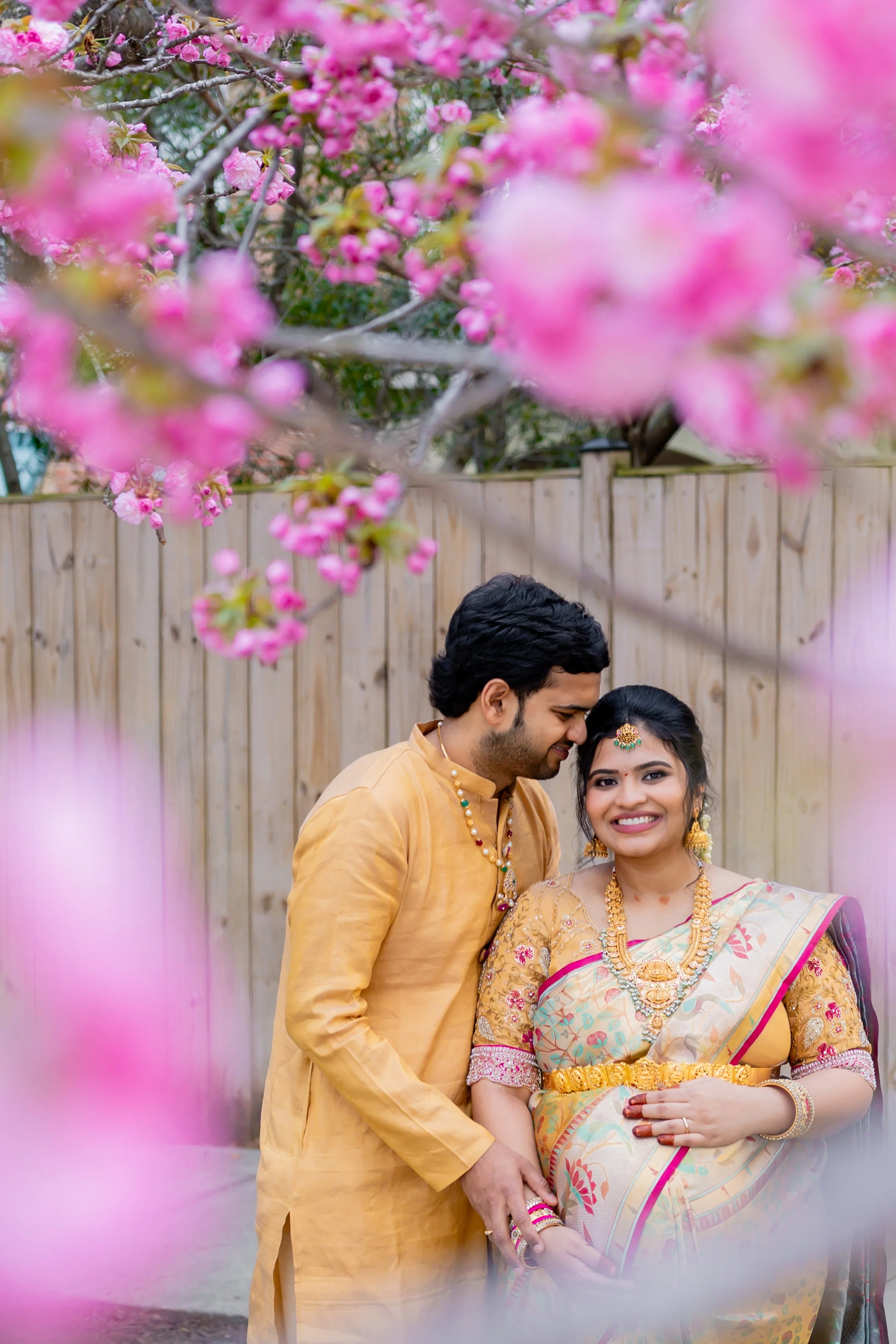 A couple dressed in traditional Indian attire, smiling and standing close together outdoors, surrounded by pink cherry blossoms.
